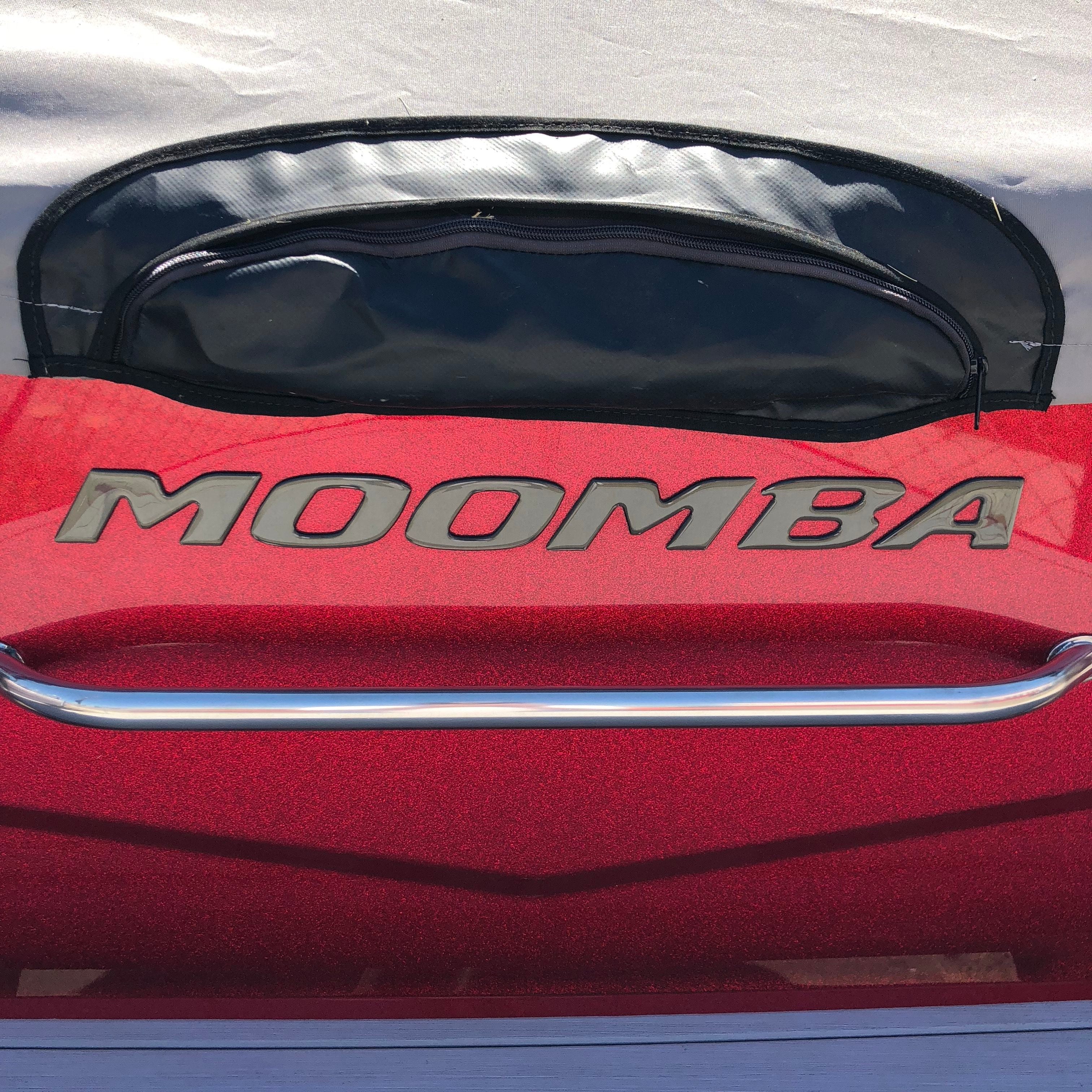 Close-up of a shiny red surface featuring the Moomba Transom Decal, Black Chrome (114193) by Moomba in large letters, a metal rail below, and part of a black and gray cover at the top of the image.