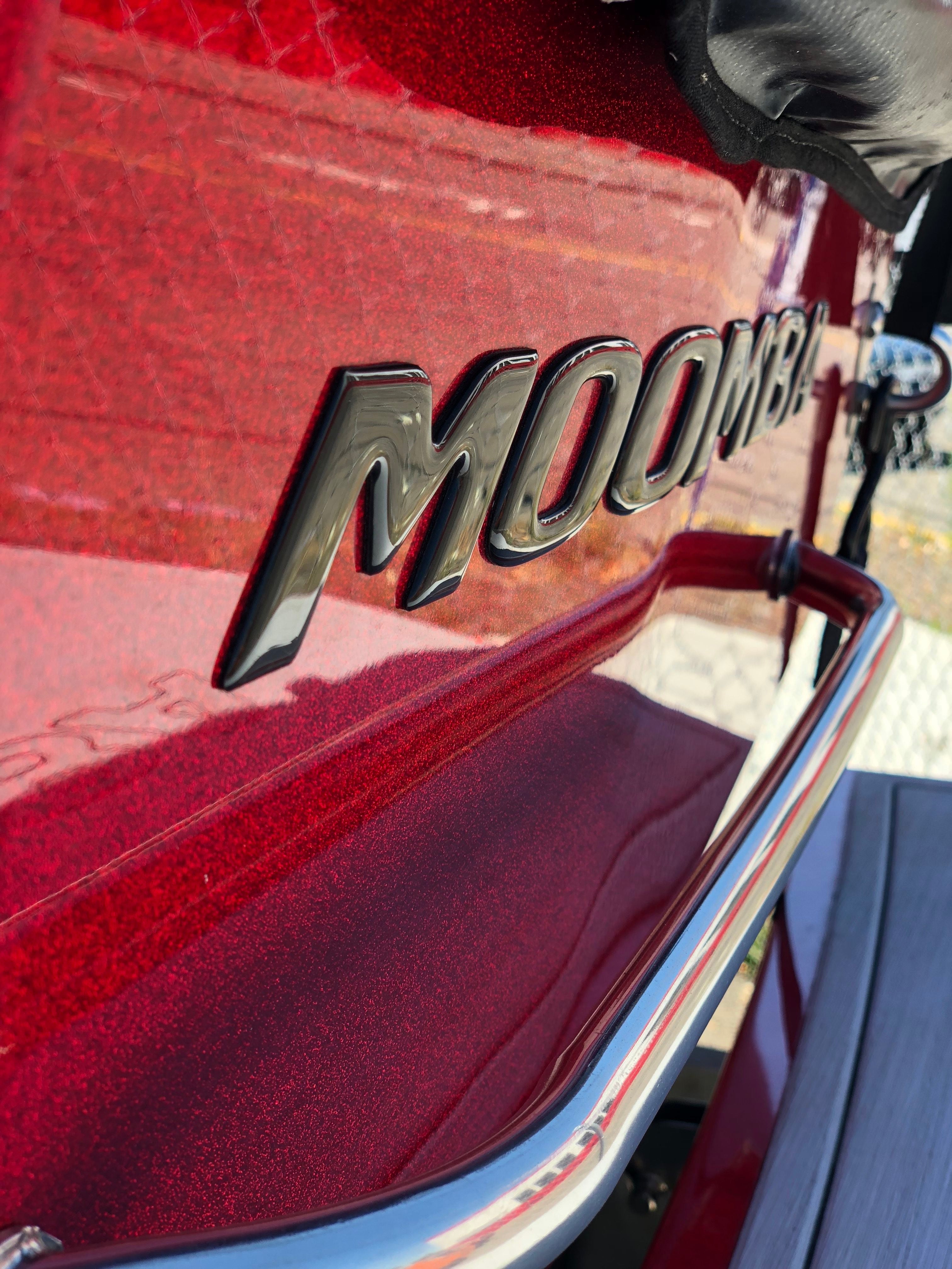 Close-up of a shiny red boat’s back with a bold Moomba Transom Decal, Black Chrome (114193) by Moomba in large black letters above a metal rail. A chain-link fence and outdoor items appear in the background.