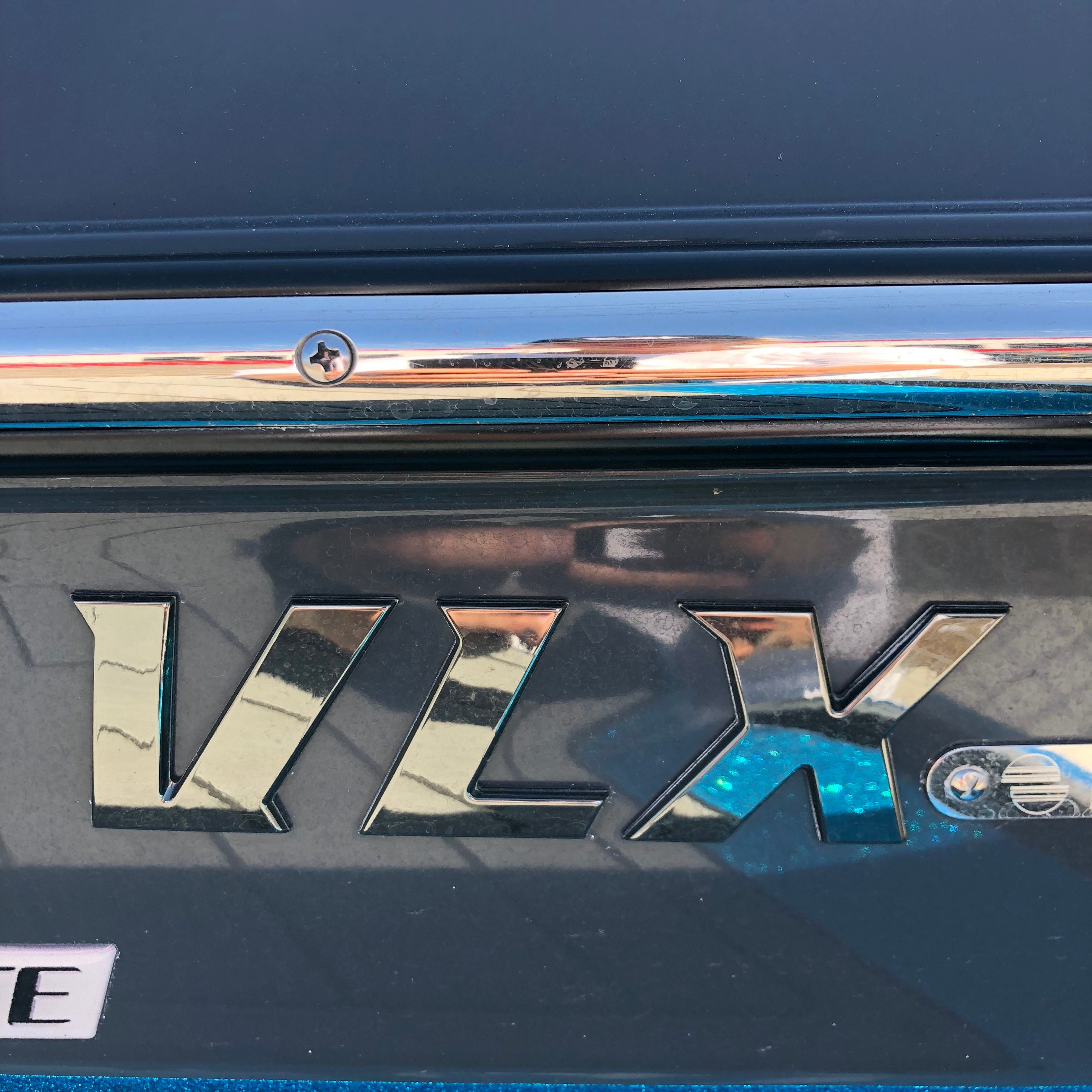 Close-up of the Malibu VLX Emblem, SL24 Black Pearl Decal (5992870) by Malibu on a blue surface, reflecting surroundings. A chrome strip with a screw runs above the emblem.