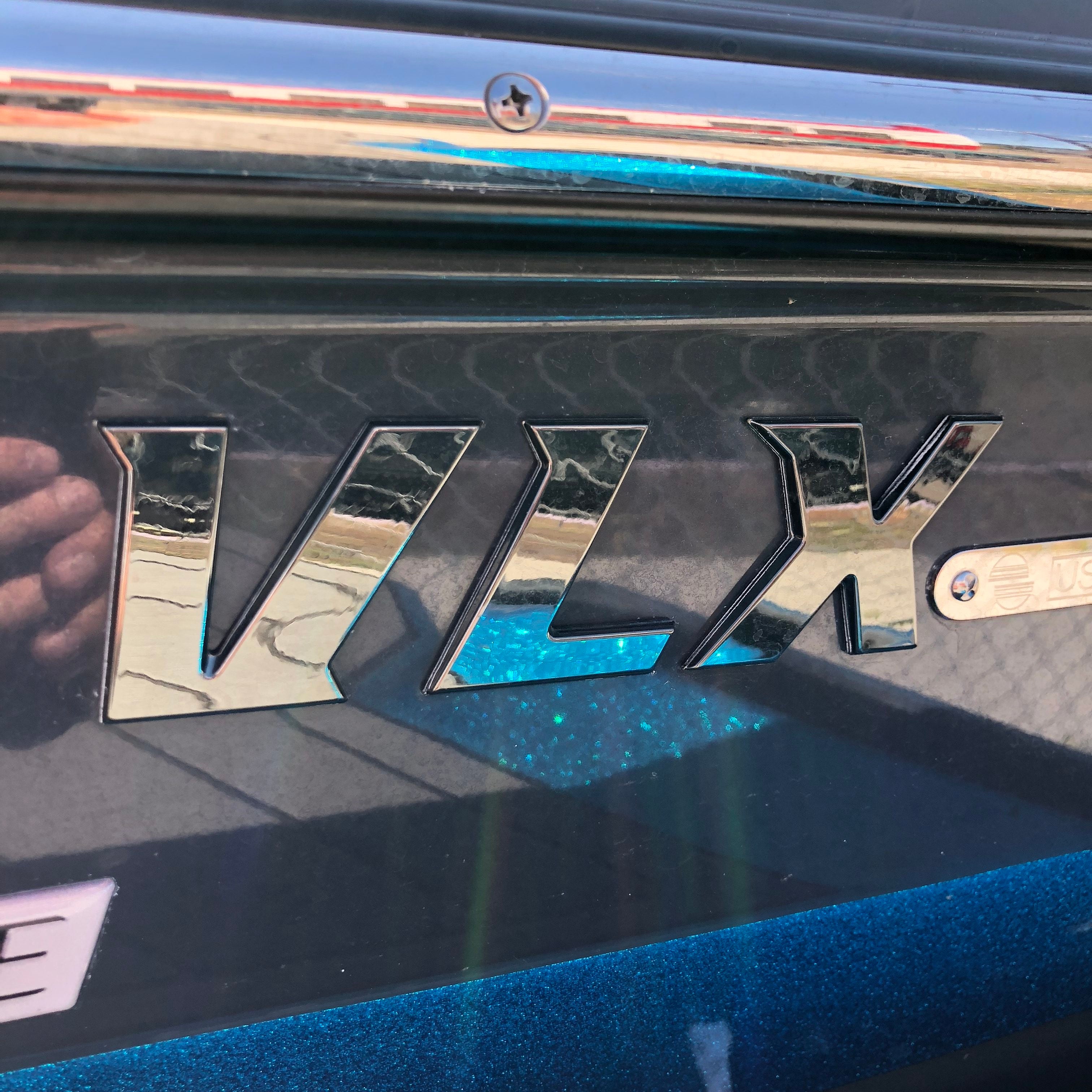 Close-up of Malibu VLX Emblem, SL24 Black Pearl, Decal (5992870) by Malibu on the side of a dark blue boat, with shiny chrome lettering reflecting outdoor scenery in the background.