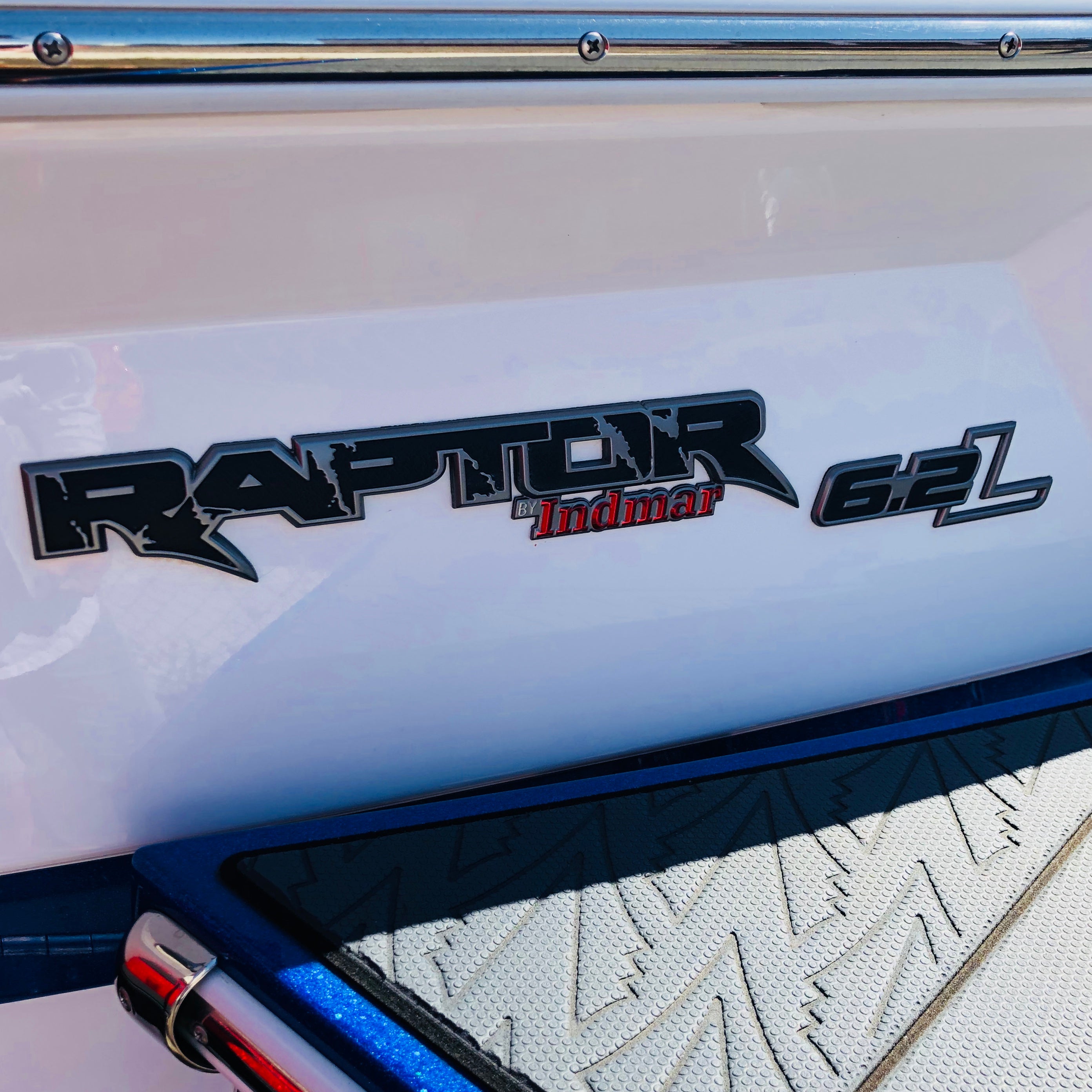 Close-up of a boat’s exterior showing the Indmar Raptor / Indmar Chromax, Badge Decal - 985417 on a white surface, with part of a textured platform and chrome trim visible.