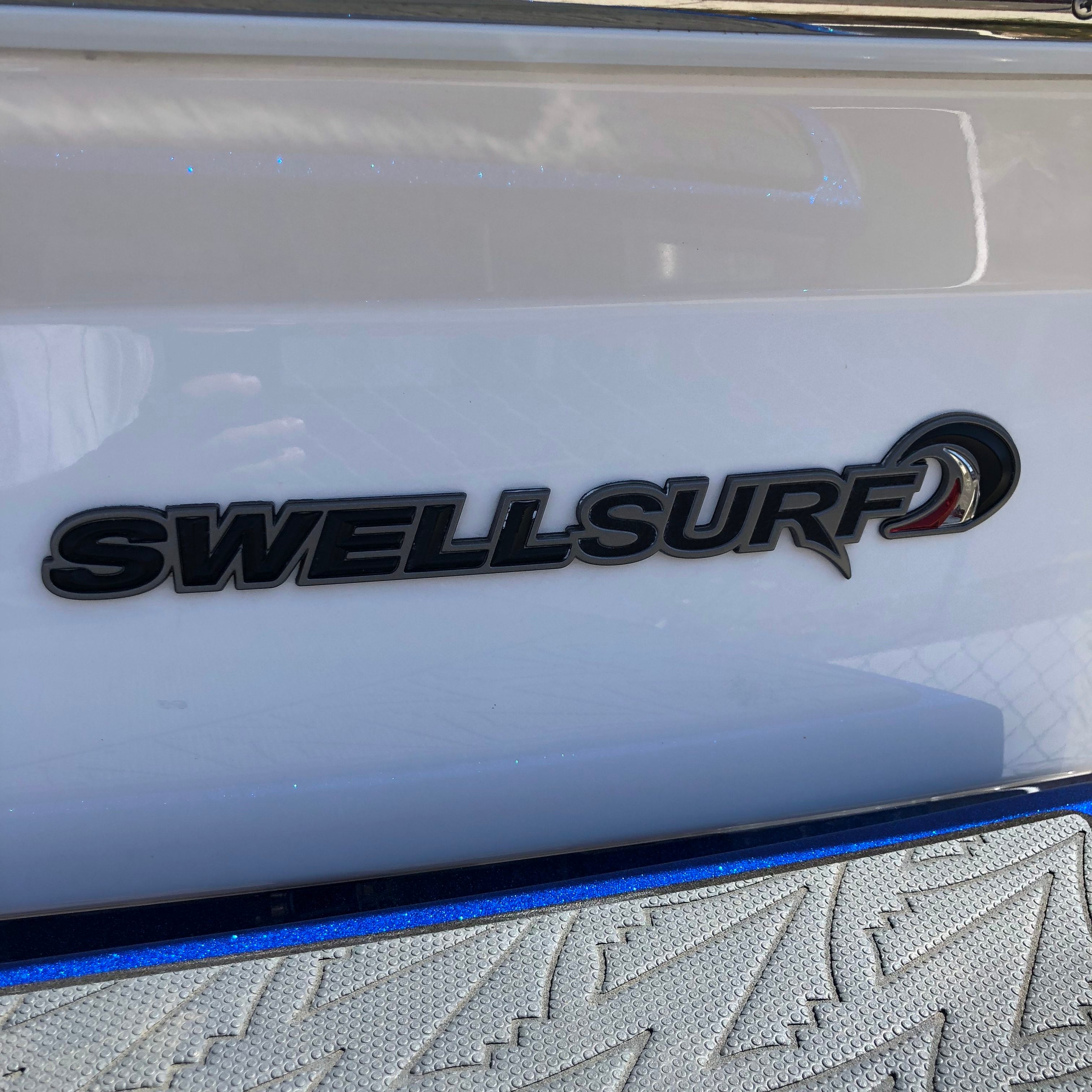 The image features the Supra Swell Surf Badge Decal by Supra in black lettering with a wave design on a glossy white surface, likely on a wakesurf boat, with a textured gray and blue mat visible at the bottom.