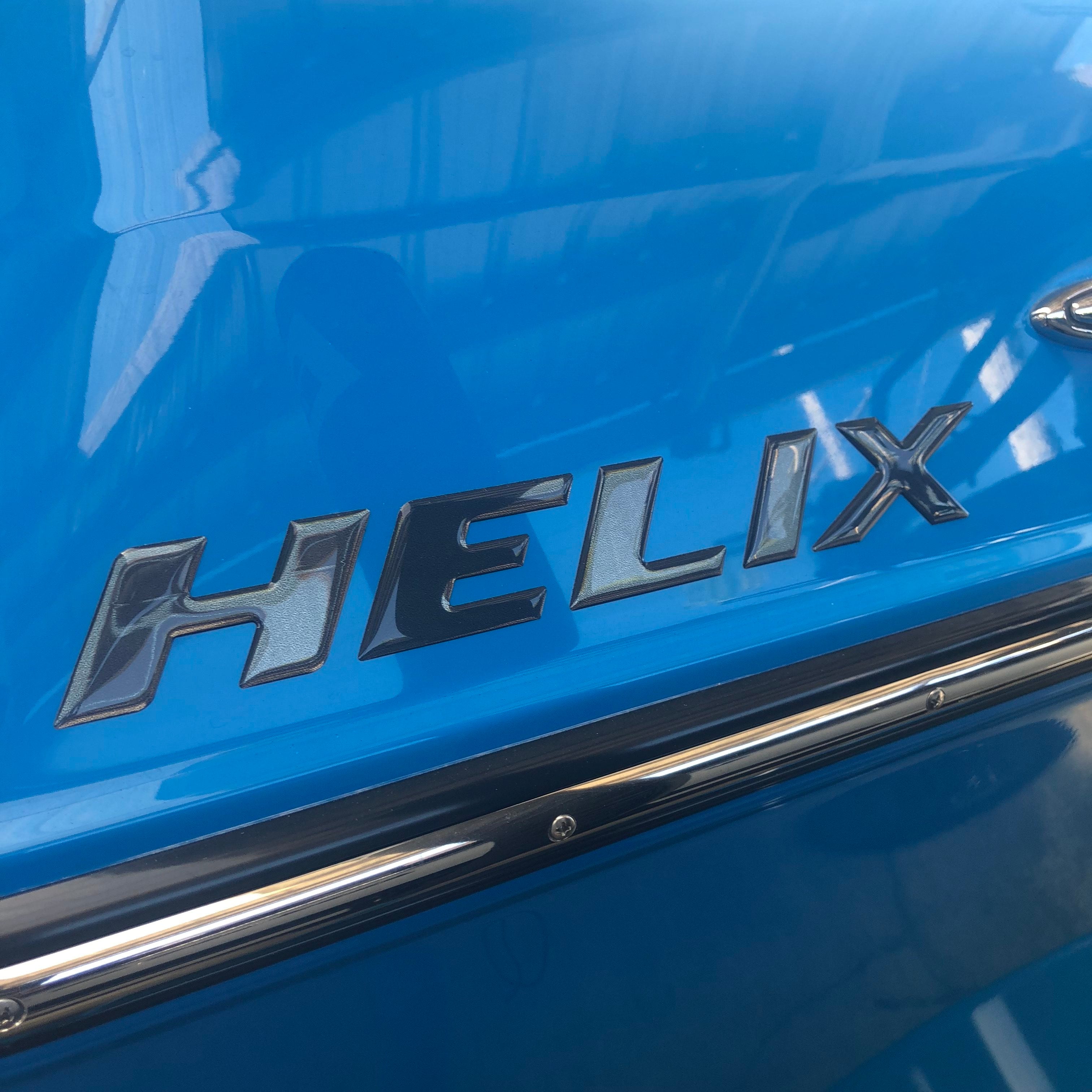 Close-up of a shiny blue Moomba with the Moomba Helix Black Chrome Designator Decal in bold black chrome. A metal trim runs below the transom decal, and reflections are visible on the paint.