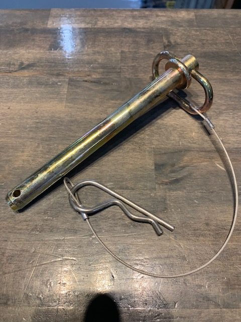The Malibu Swing Tongue Pin 6.5" with locking ring and wire is attached to a bent metal hitch pin clip, positioned on a wooden trailer surface.