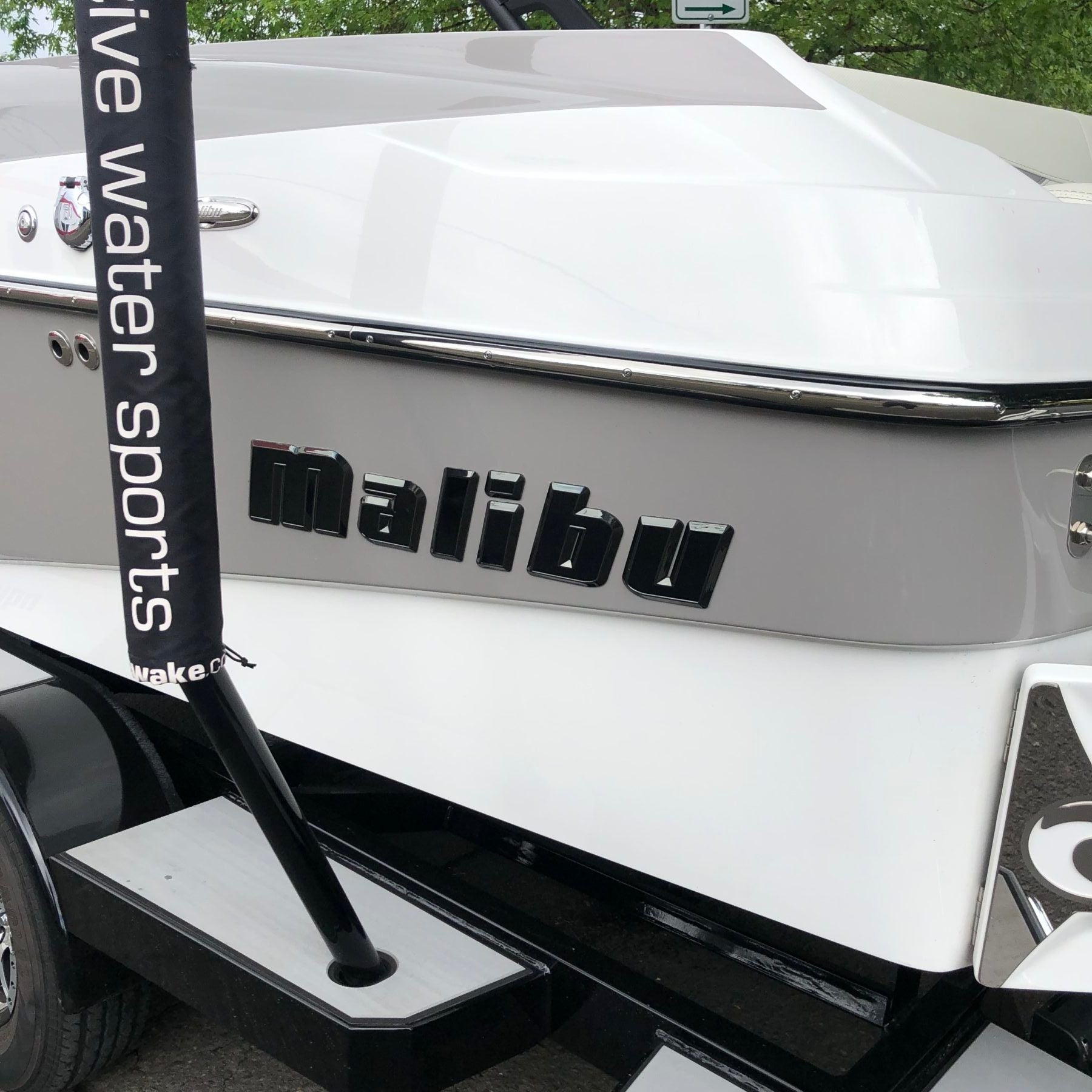 A close-up of the back of a Malibu boat displays the Malibu Hull Decal, 26" Black Pearl, Black Chrome, SL24 by Malibu. Part of a black pole with “live water sports” text is also visible.