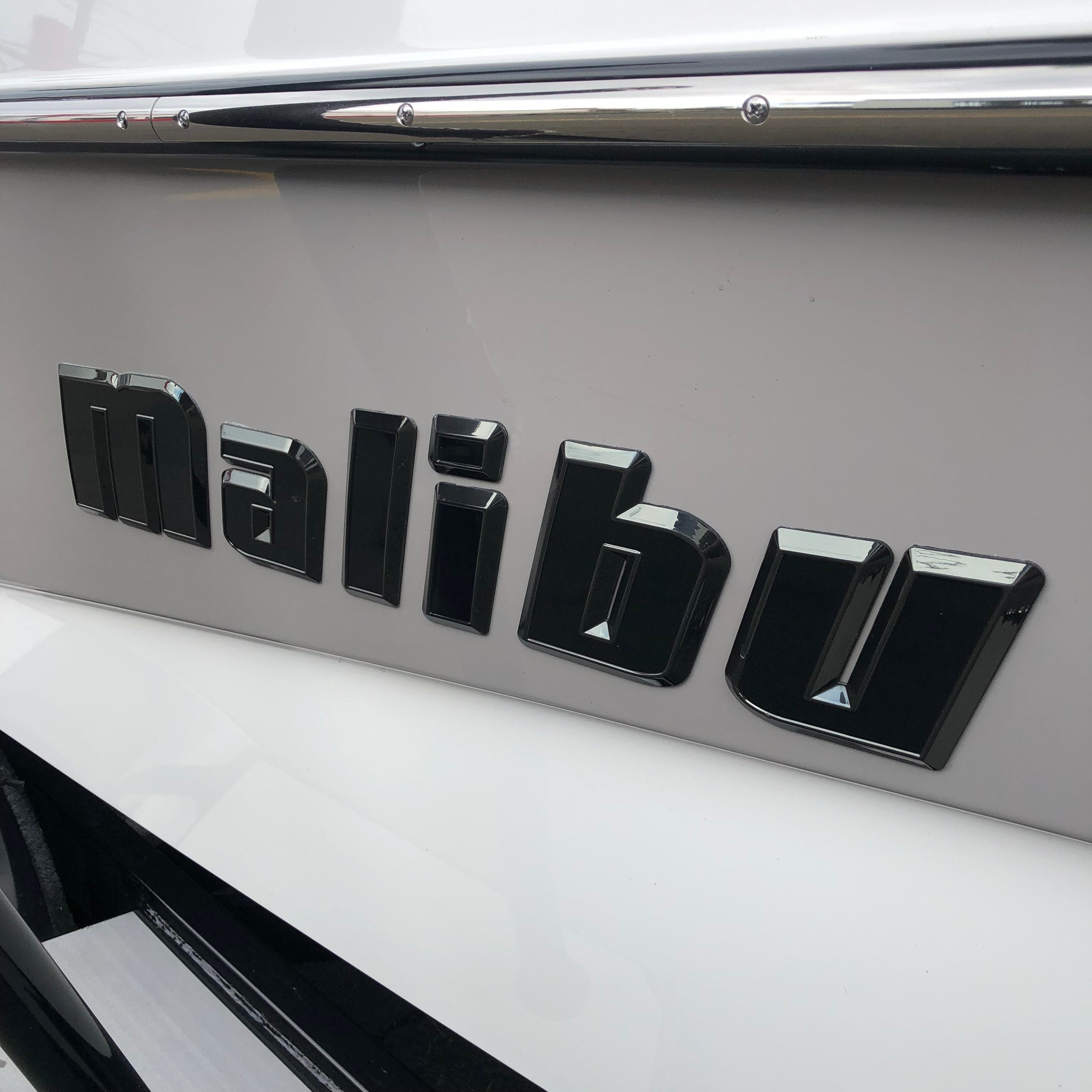 Close-up of the Malibu Hull Decal, 26" Black Pearl, Black Chrome (SL24) by Malibu displayed on the side of a white boat, with chrome trim visible above the emblem.