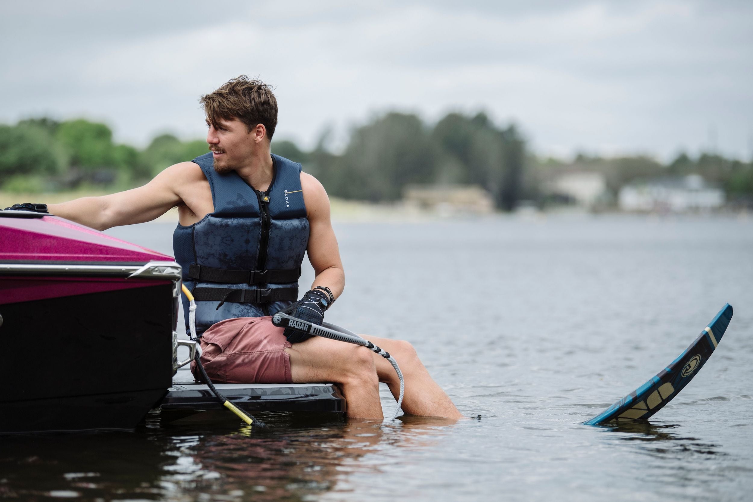 Wearing a life vest, a man sits at the back of the boat, firmly holding onto the Radar Control Barlock Pro Package handle from Radar. With one foot secured in a water ski, he gets ready for an exciting adventure on the serene lake.