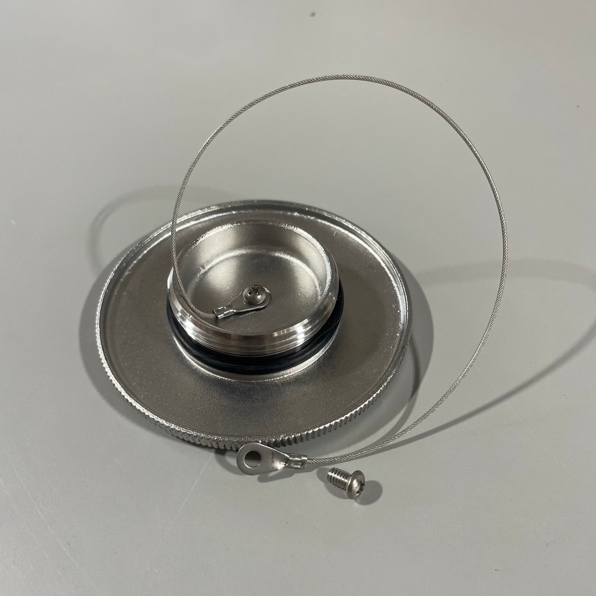 The Malibu Gas Cap 9-11 Replacement by Malibu features a large chrome cap with a screw, metal loop, and attached cable, designed for secure lid locking. It is shown resting on a light-colored surface.