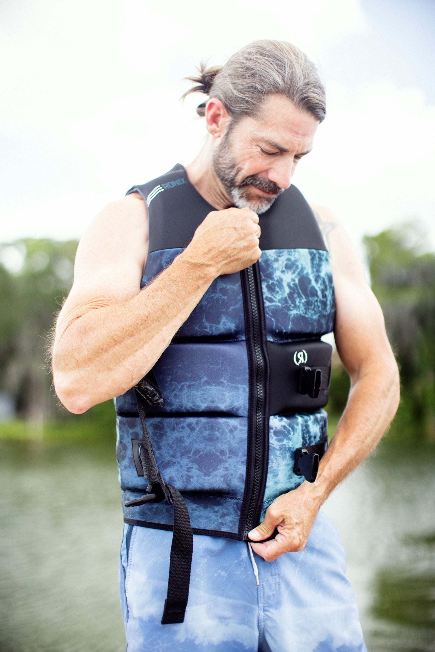 A man wearing a Ronix 2024 Point Break Yes Men's CGA Vest standing next to a body of water, prepared for any potential sea dwelling emergency.