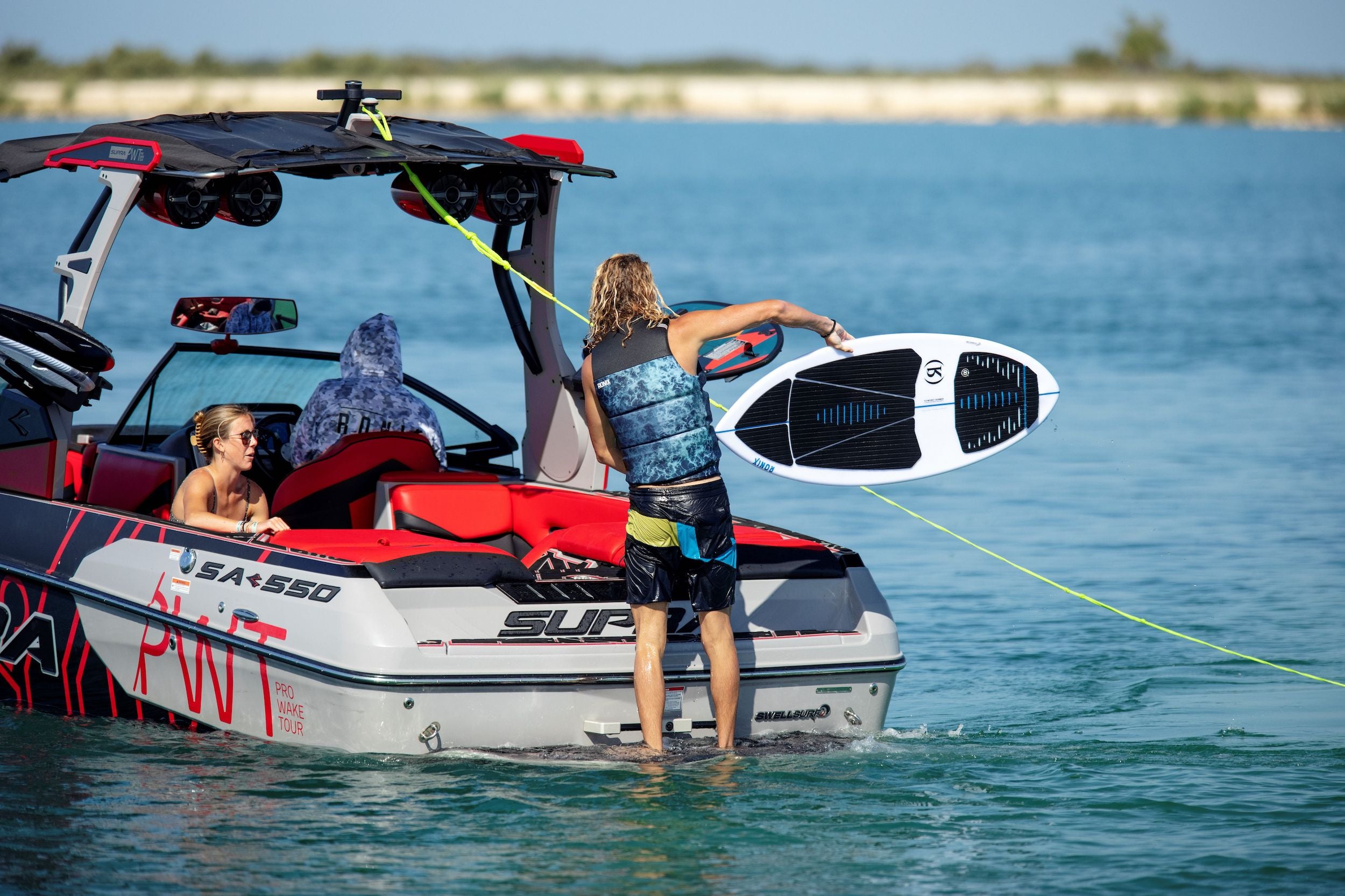 A man, wearing a Ronix 2024 Point Break Yes Men's CGA Vest, is standing on the back of a boat with a wakeboard.