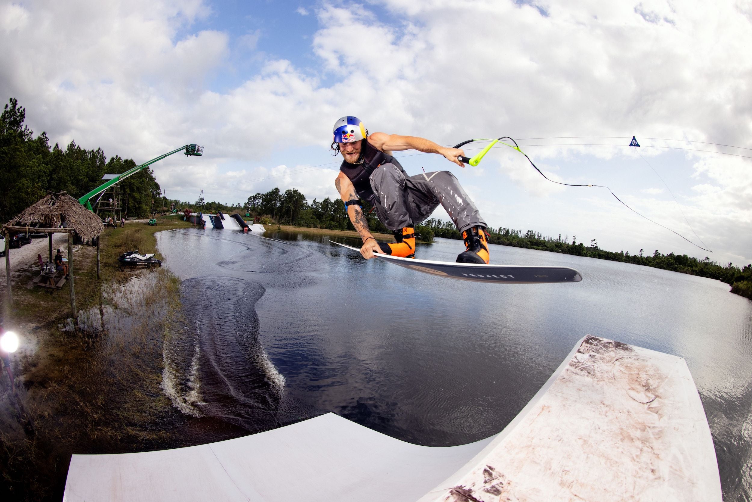 A wakeboarder in a helmet and vest catches air above a ramp, gripping the cable while wearing Ronix 2023 RXT Boa Boots with a heat moldable liner, set against a scenic outdoor backdrop and blue sky.