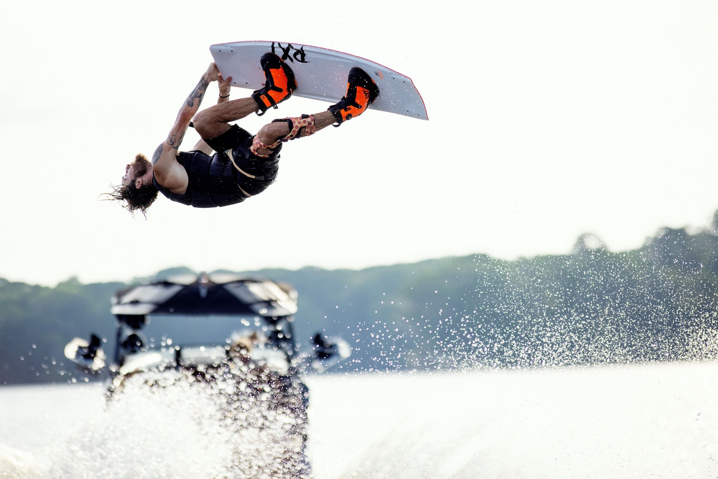 Wearing Ronix 2023 RXT Boa Boots, a wakeboarder performs an aerial flip above the water while being towed by a boat, with water spray and distant trees in the background.