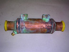 An Indmar COOLER 5" STD w/ORING THREAD - 865003 heat exchanger by Indmar, with green oxidation, yellow-capped threaded ends, and small top fittings, is shown on a plain surface.