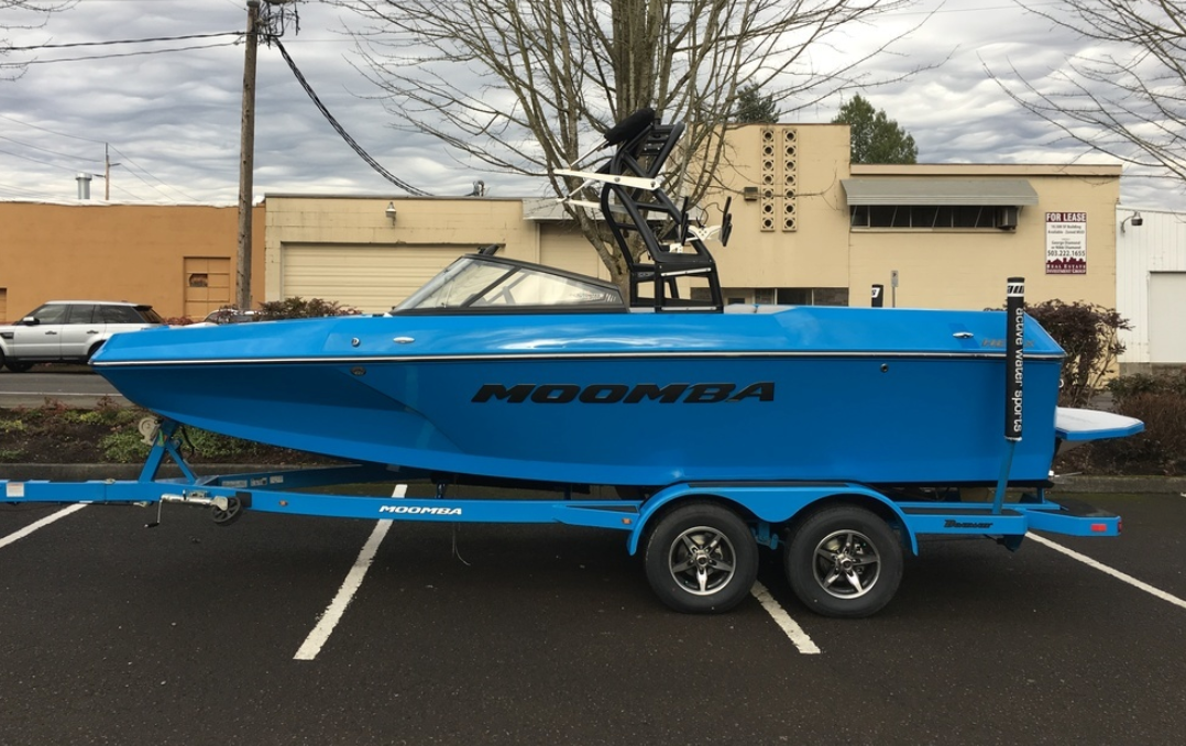 A bright blue Moomba speedboat featuring the Moomba Helix Black Chrome Designator Decal is parked on a matching blue trailer in a lot, with buildings and leafless trees in the background under a cloudy sky.