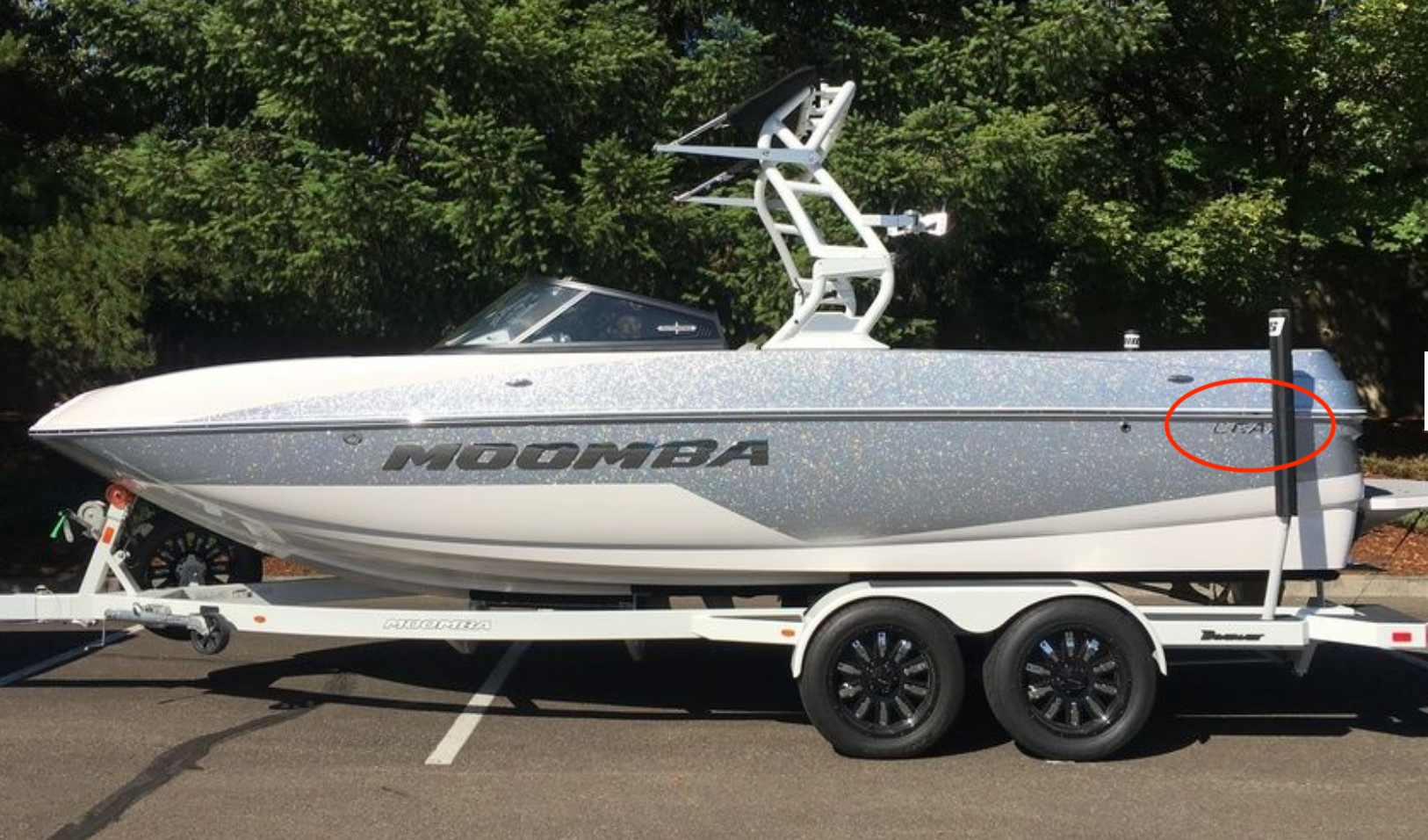A Moomba CRAZ speedboat, featuring the Moomba CRAZ Chrome Transom Decal (116054) from Moomba, is parked on a trailer in a wooded lot. The chrome decal and a registration sticker are visible on the boat's side near the back.
