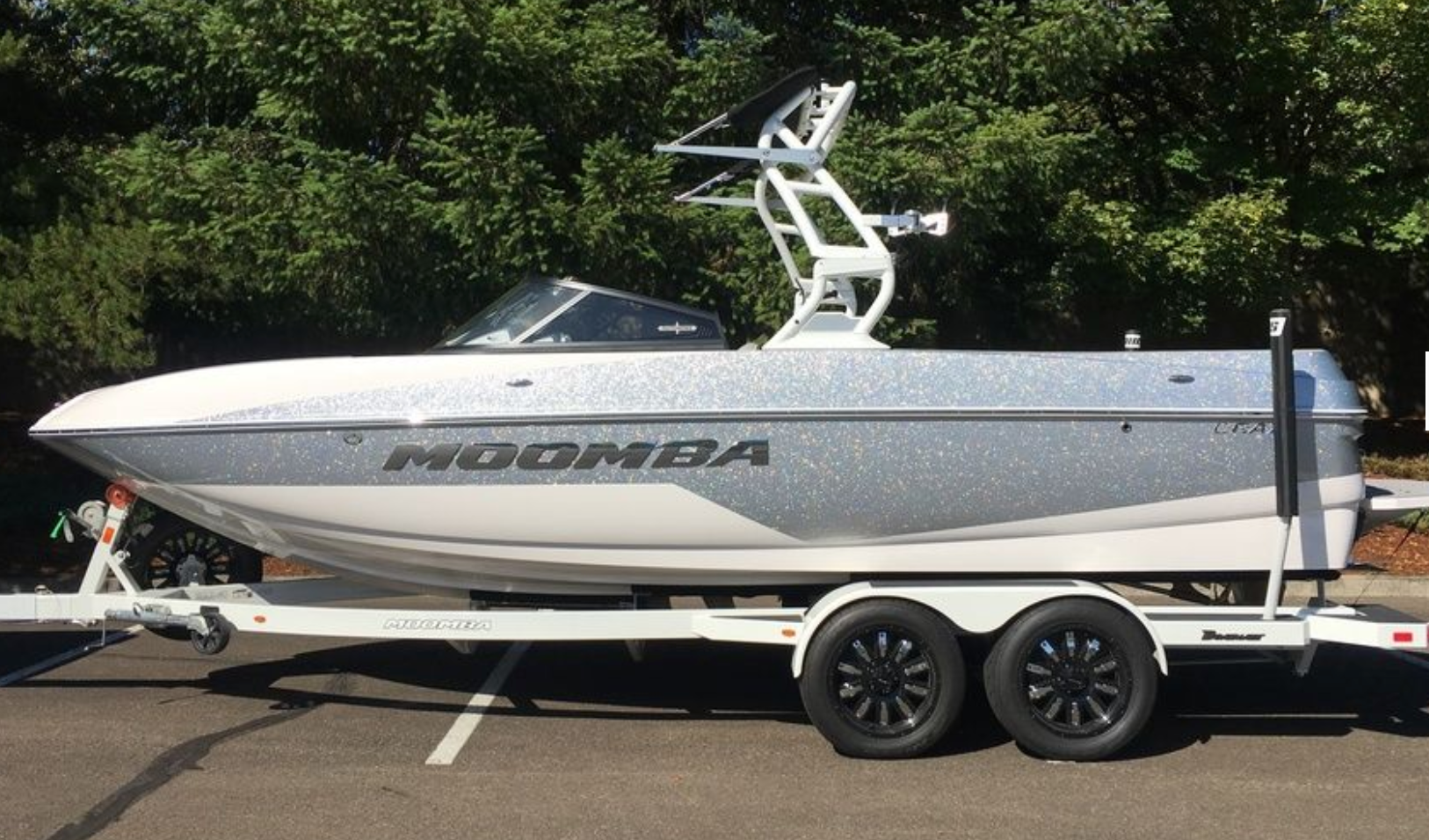 A Moomba boat featuring the Moomba Chrome Badge, Hull Decal (110150) by Moomba sits on a double-axle trailer in a parking lot, surrounded by trees.