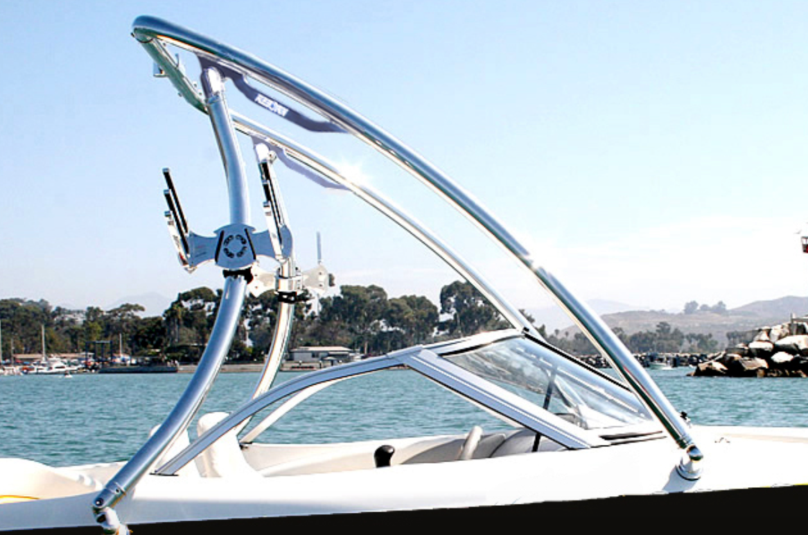 A close-up of the Reborn Elevate Wakeboard Tower Shining Polished by Reborn, mounted on a white boat with clear blue water and trees in the background under a bright sky.