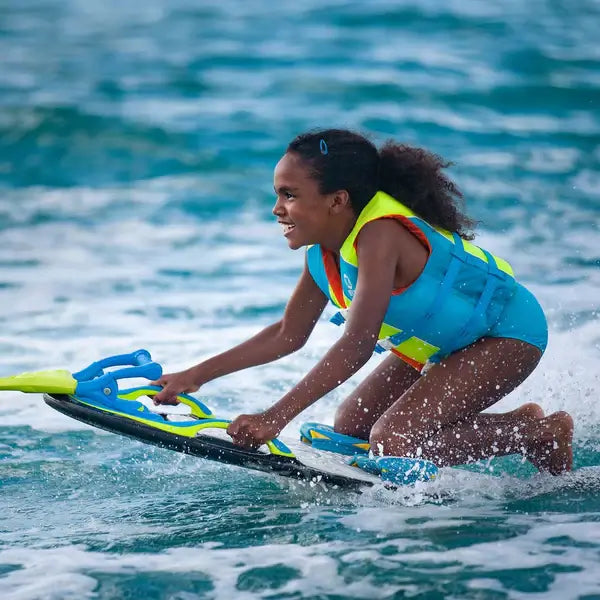 A young girl in a blue life jacket smiles as she rides the ZUP YouGotThis 2.0 Board by ZUP, gliding over clear blue water on this stable, beginner-friendly board with a hydrodynamic design.