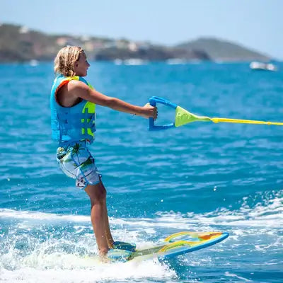 A person in a blue life jacket and patterned shorts is wakeboarding on bright blue water with the ZUP YouGotThis 2.0 Board - Black by ZUP, holding a tow rope, with a hilly coastline visible in the background.