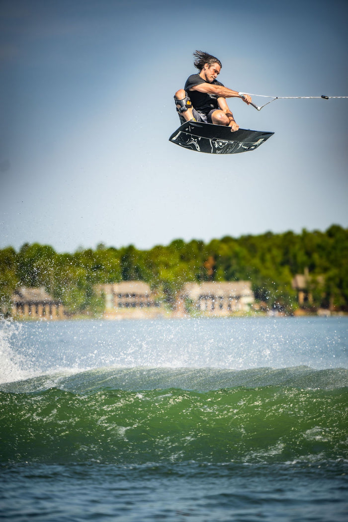 A man wakeboarding in the air over a lake, showcasing his skills with the Hyperlite 2023 Cryptic Wakeboard and Team OT Bindings.