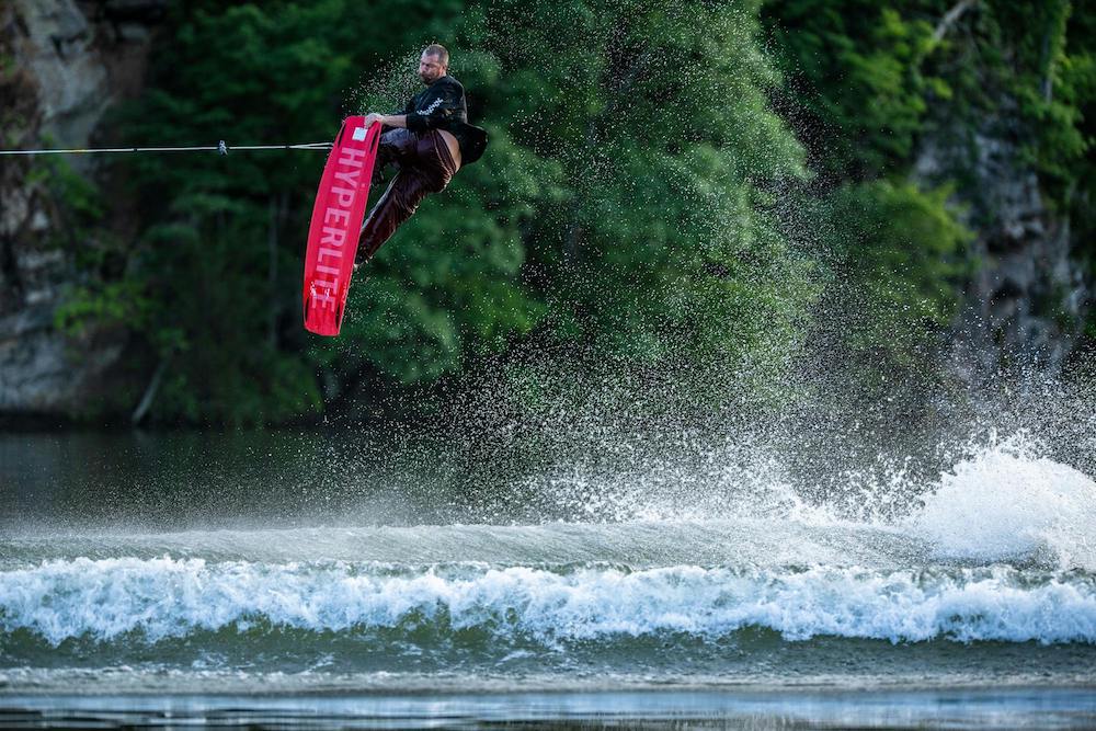 A person wakeboarding pops off the wake, performing a jump over water while holding a rope. The Hyperlite 2025 Blueprint Wakeboard, known for delivering the softest landings, is featured. Lush vegetation is visible in the background.