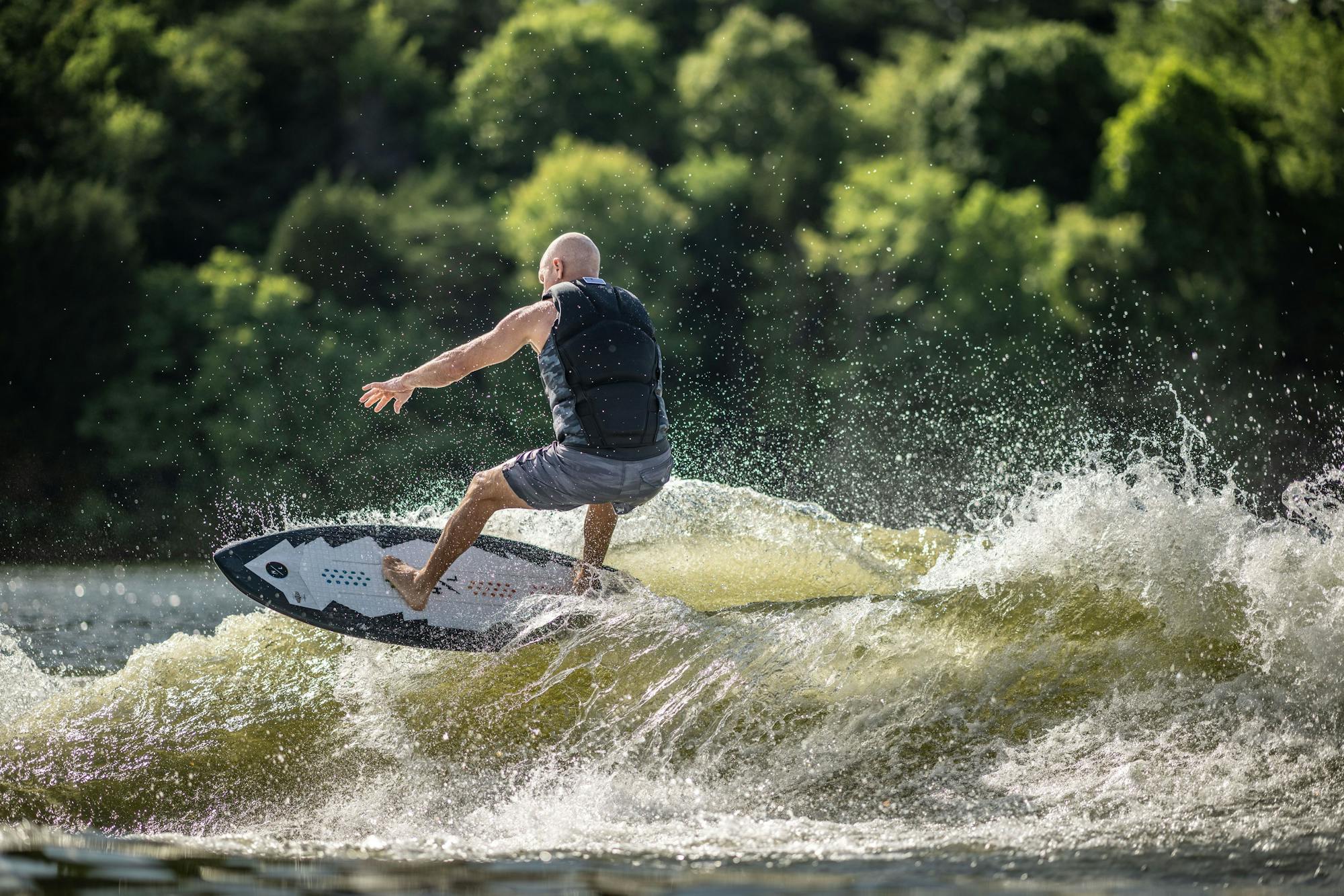 A man wearing a life vest is wakesurfing on a Hyperlite 2025 Broadcast Wakesurf Board, effortlessly gliding through splashing waves on the lake, with trees framing the picturesque scene.