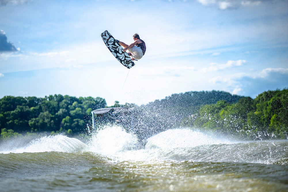 A person riding a Hyperlite 2025 Capitol Wakeboard, enhanced by the Blended 3-Stage Rocker, performs a mid-air trick above the shimmering water, framed by vibrant trees and a sunny sky.