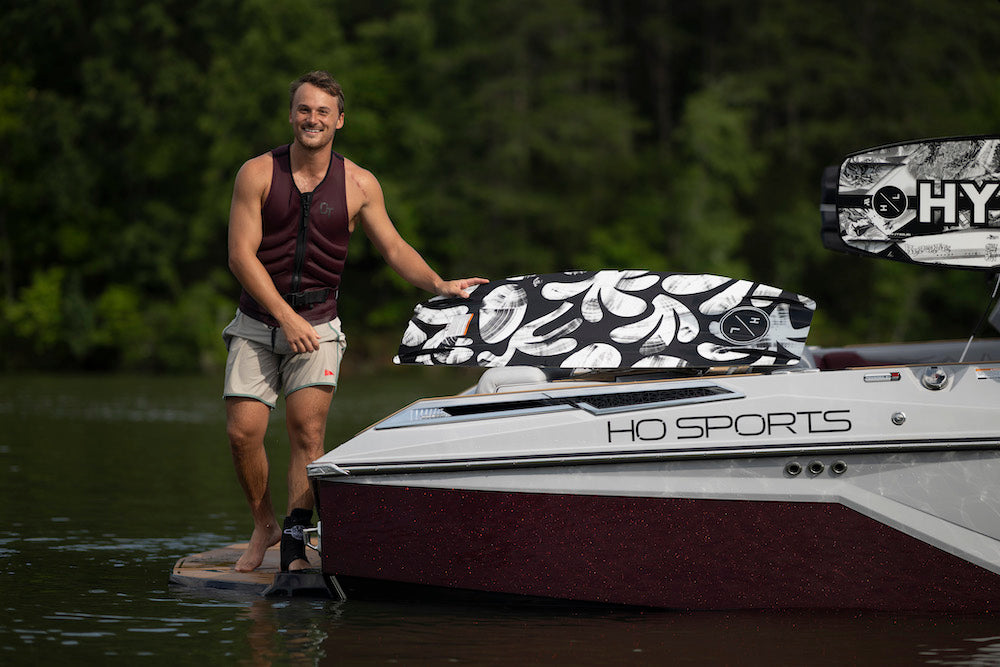 A man in a sleeveless vest stands on a dock beside a boat, with a Hyperlite 2025 Capitol Wakeboard featuring a Biolite 3 Core resting nearby. Trees and serene water create the perfect backdrop for this action-packed scene.