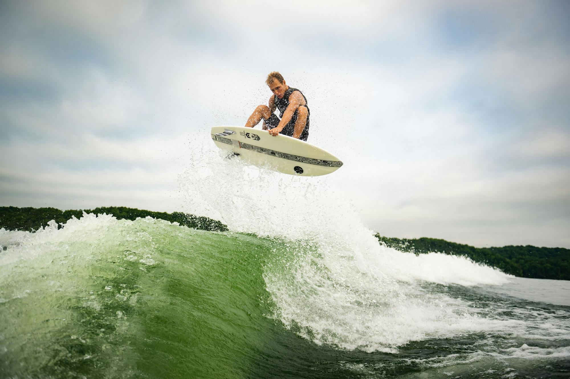 A wakesurfer skillfully executes an aerial maneuver on the Hyperlite 2025 Condor Wakesurf Board, slicing through the water beneath a cloudy sky.