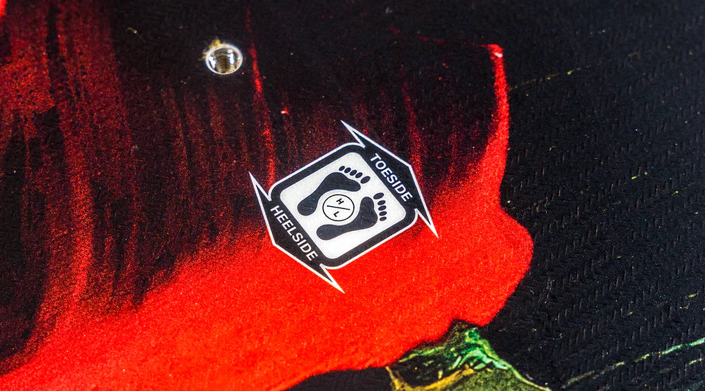 Close-up of an asymmetrical design on a black surface with red and green elements, featuring a sticker illustrating foot placement directions labeled toeside and heelside, perfect for mastering toe-side jumps on your Hyperlite 2025 Eden Jr Wakeboard.