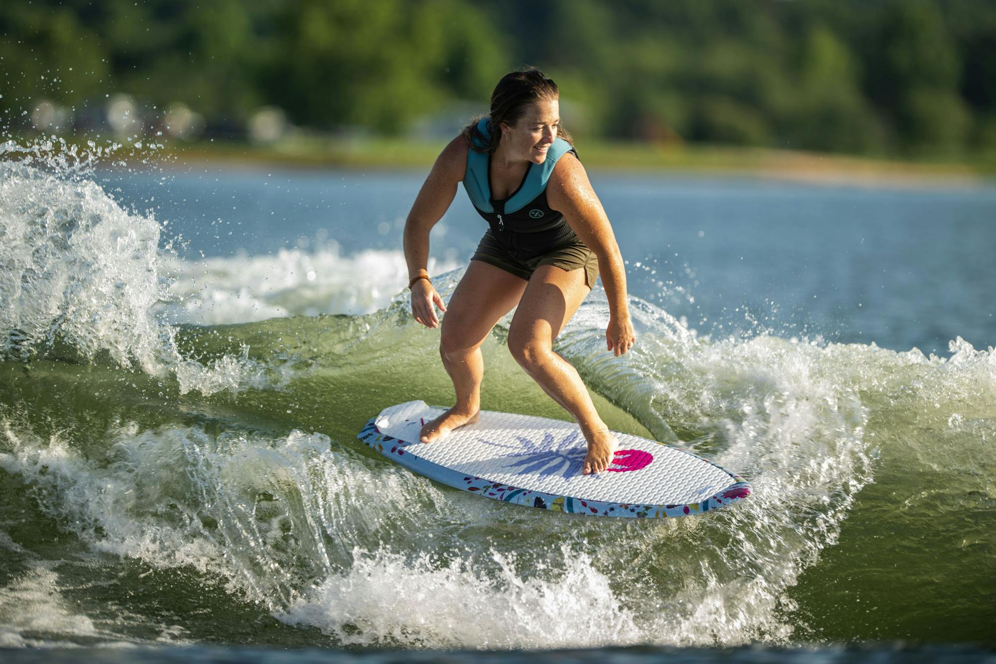 A wakesurf enthusiast effortlessly rides the waves in a lake, outfitted with a life jacket and shorts. It's just another day of catching waves on the Hyperlite 2025 Good Daze Women's Wakesurf Board.