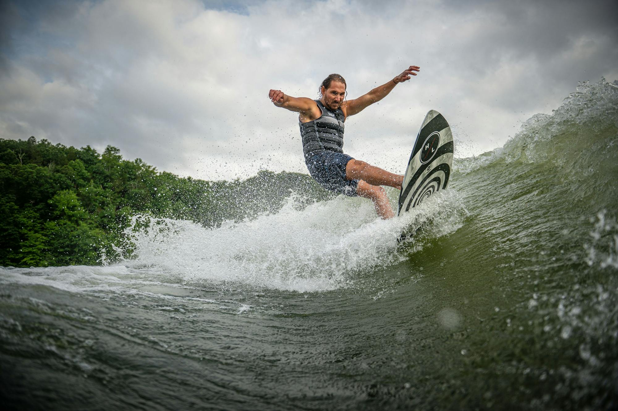 Using the Hyperlite 2025 Hi-Fi Wakesurf Board, a person in a life vest expertly surfs on a wave, their balance enhanced by an EVA traction pad. They are enveloped by lush greenery and a cloudy sky, illustrating nature's mix of tranquility and untamed energy.