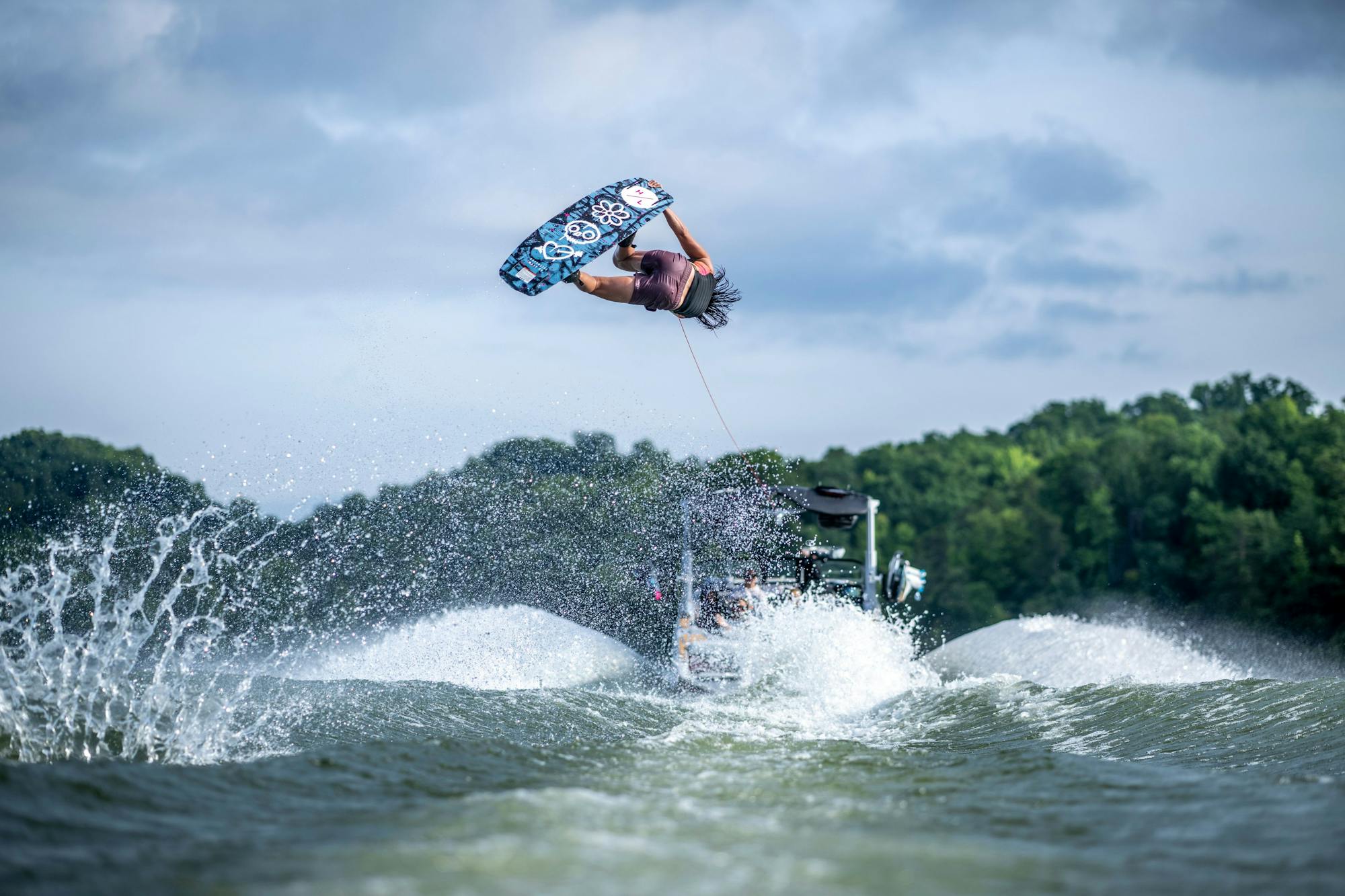 A person performs an aerial flip with precision while wakeboarding on the Hyperlite 2025 Journey Wakeboard, featuring a 3-Stage Rocker, behind a boat gliding over the lake.