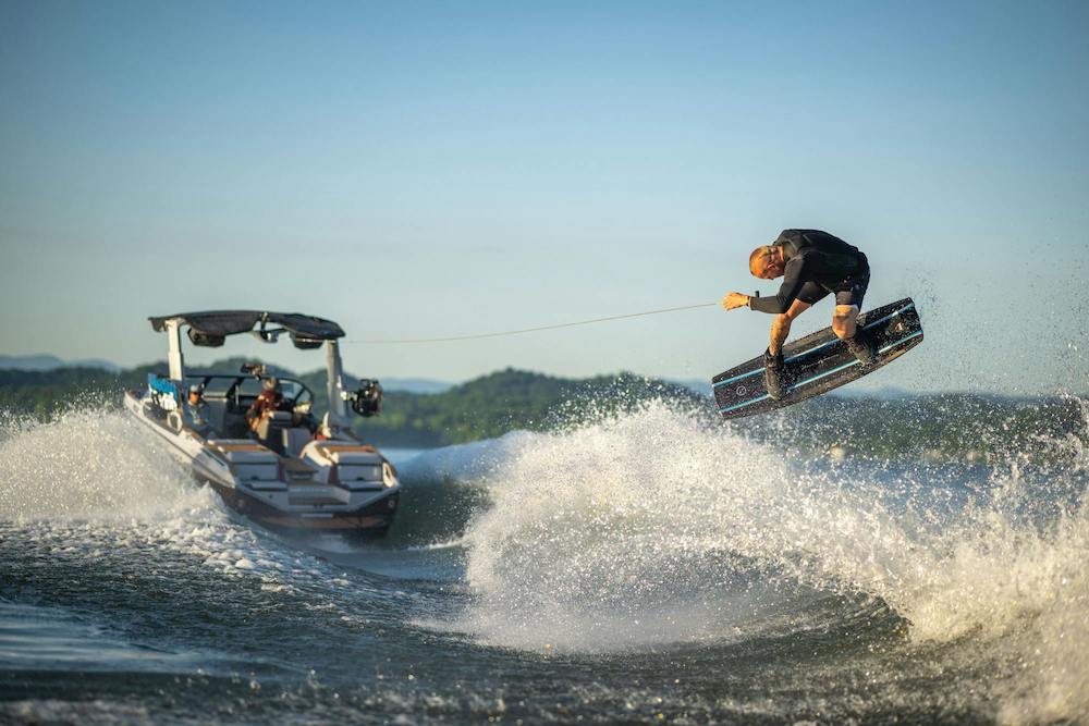 A person is wakeboarding on a lake, performing a jump while skillfully executing a Boneline Grab Rail maneuver using the Hyperlite 2025 Kruzr-GLS Wakeboard. Holding the rope attached to the motorboat, they soar through the air as the boat speeds ahead, sending splashes across the water's surface.