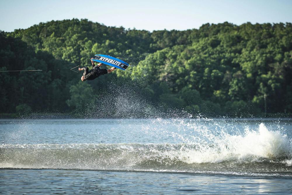 A person on a wakeboard outfitted with a Hyperlite 2025 Kruzr-GLS performs a flip above the lake, surrounded by trees and splashes of water below.