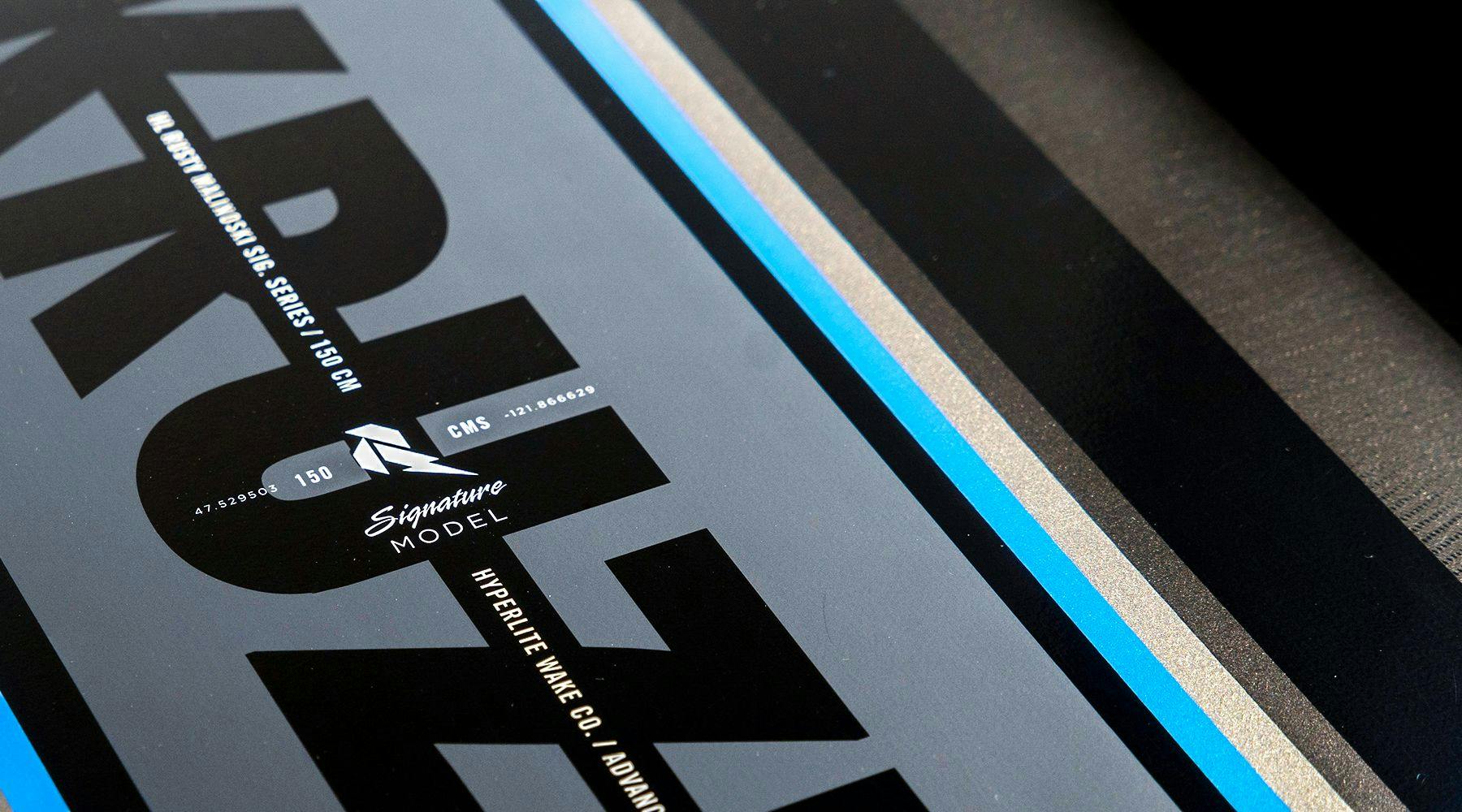 Close-up of the Hyperlite 2025 Kruzr-GLS Wakeboard displaying the text Signature Model, showcasing a black, blue, and gray design with Boneline Grab Rail for improved performance.