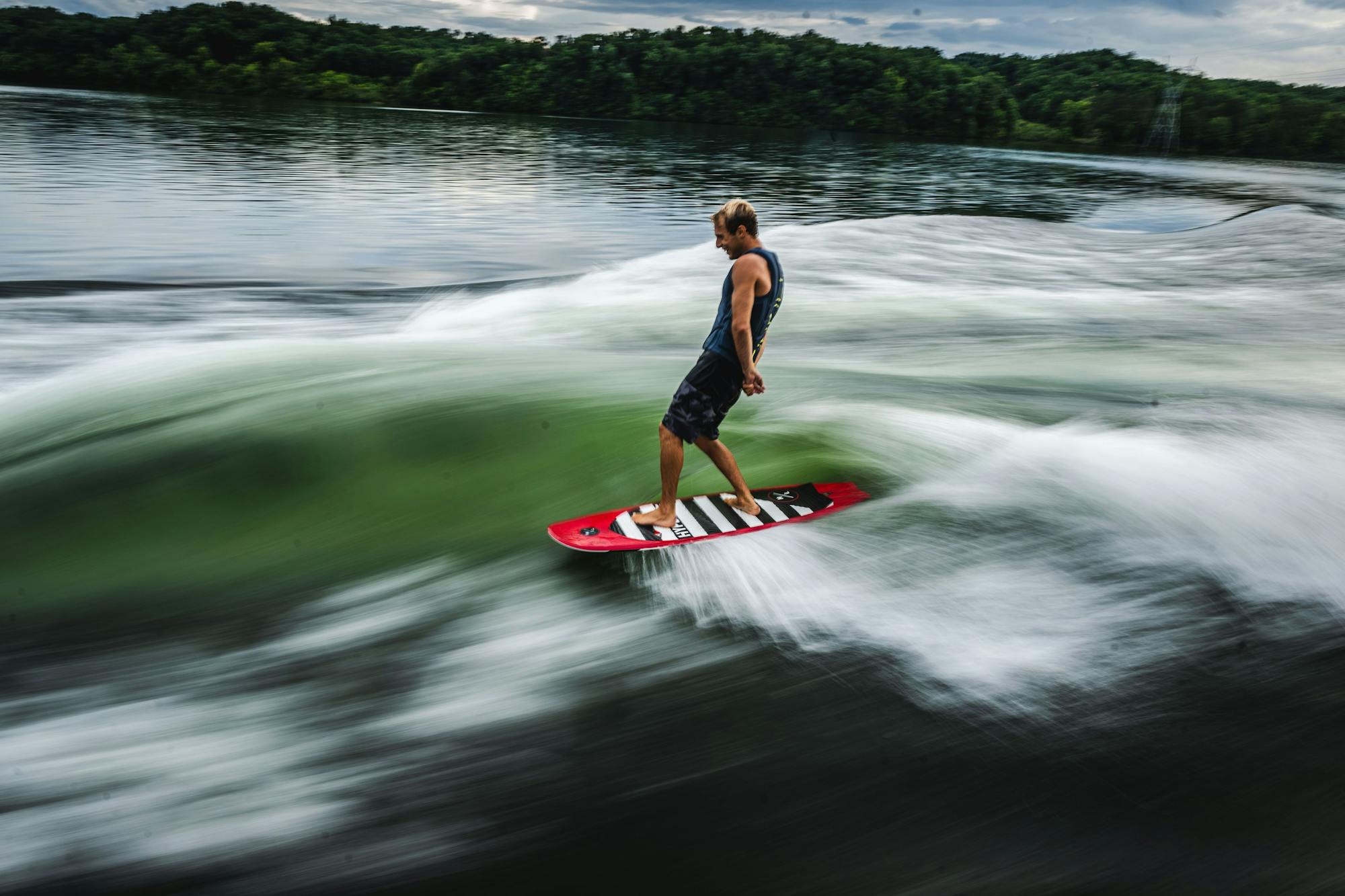 A surfer showcases their style on a Hyperlite 2025 Landlock Wakesurf Board - 5'9" as they ride a wave in the water, with a forested shoreline creating a beautiful backdrop.