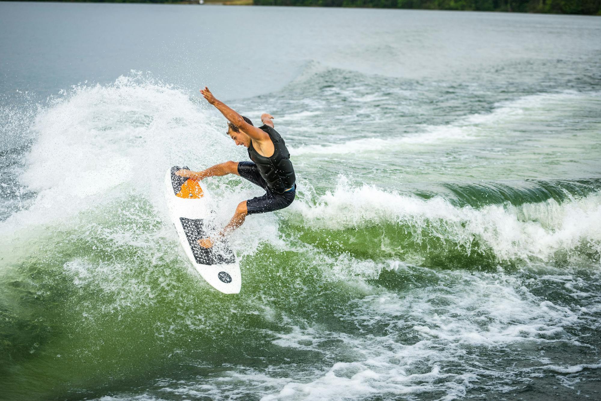 A person gliding across a wave, performing an aerial maneuver on the Hyperlite 2025 Lobo Wakesurf Board, while wearing a black wetsuit and surrounded by the spray of water.