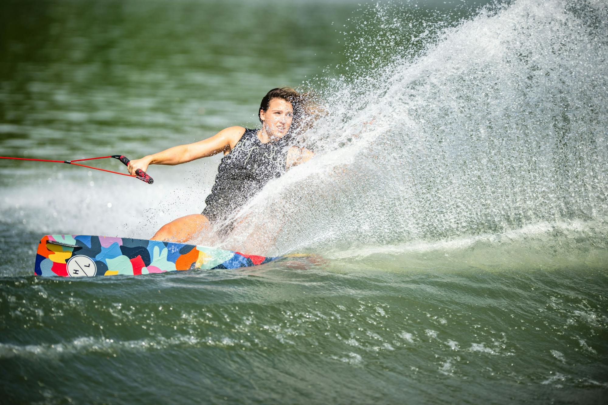 A person skillfully wakeboarding on a colorful Hyperlite 2025 Prizm Wakeboard, equipped with a subtle 3-Stage Rocker, creates a large spray of water.