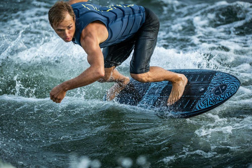 A person in a blue vest and black shorts is wakesurfing on the Hyperlite 2025 Ranger Wakesurf Board, expertly demonstrating surf style as they glide through choppy water.