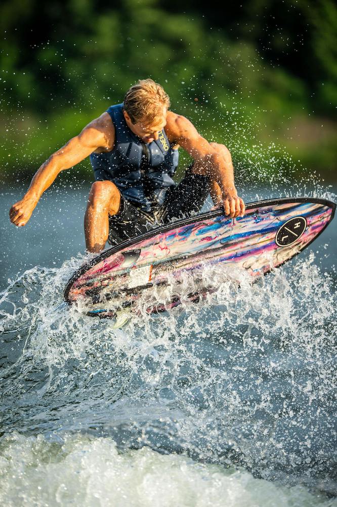 A person wakesurfs on a vibrant Hyperlite 2025 Ranger Wakesurf Board with DuraShell Construction, surrounded by splashing water against a backdrop of greenery.