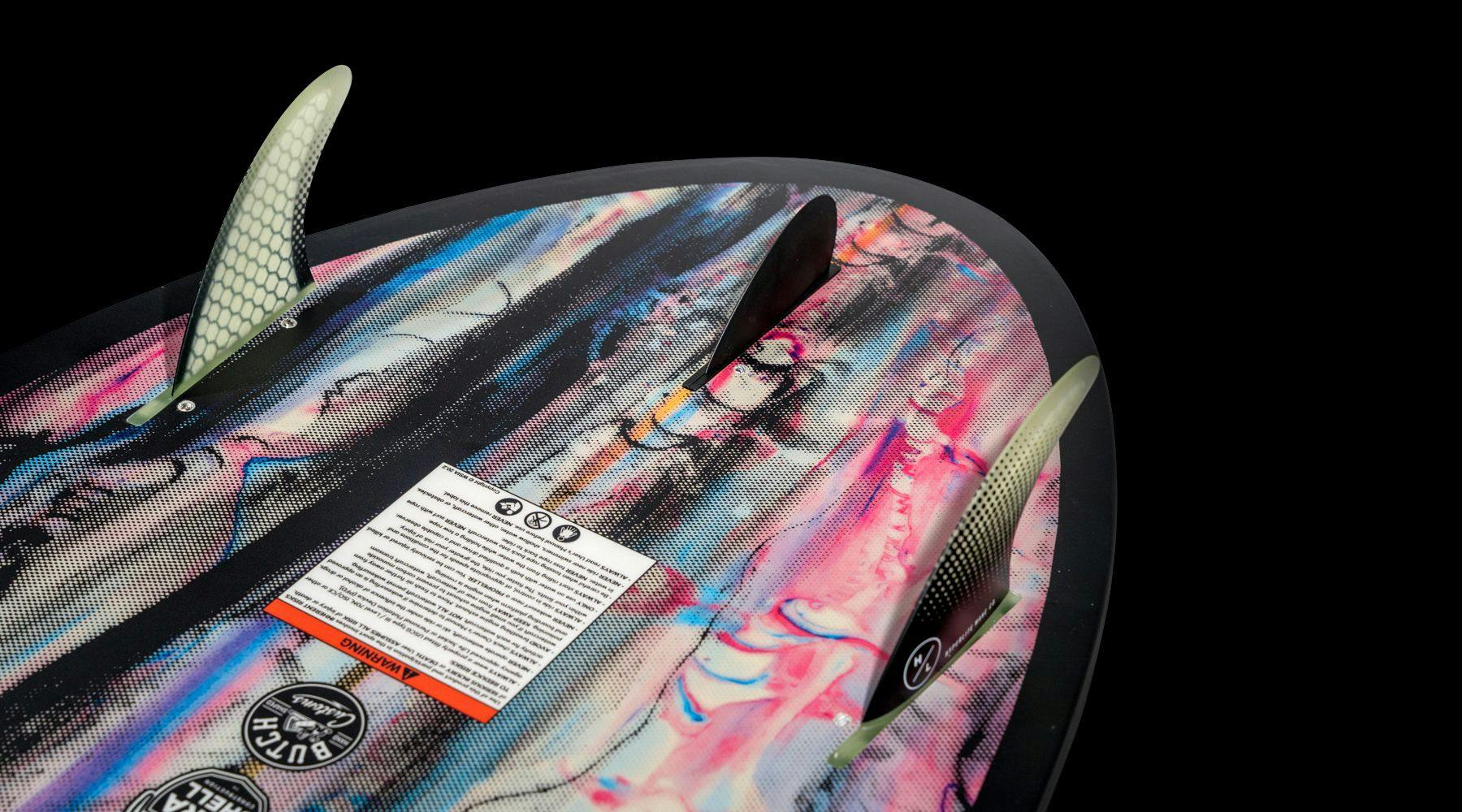 Close-up of the tail of a Hyperlite 2025 Ranger Wakesurf Board highlighting its Thruster Fin Configuration. The board features a vibrant, abstract design crafted with DuraShell Construction and includes an information label on its deck.