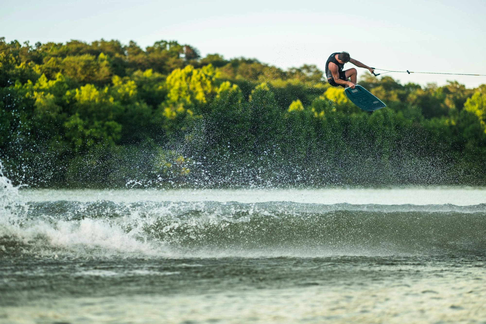 A person wakeboarding in mid-air above a shimmering lake captures the thrill of adventure, with a forested shoreline providing a stunning backdrop. The rider expertly maneuvers their Hyperlite 2025 Sender Wakeboard, enhanced by its Dual Concave Base, ensuring smooth landings and epic airtime.