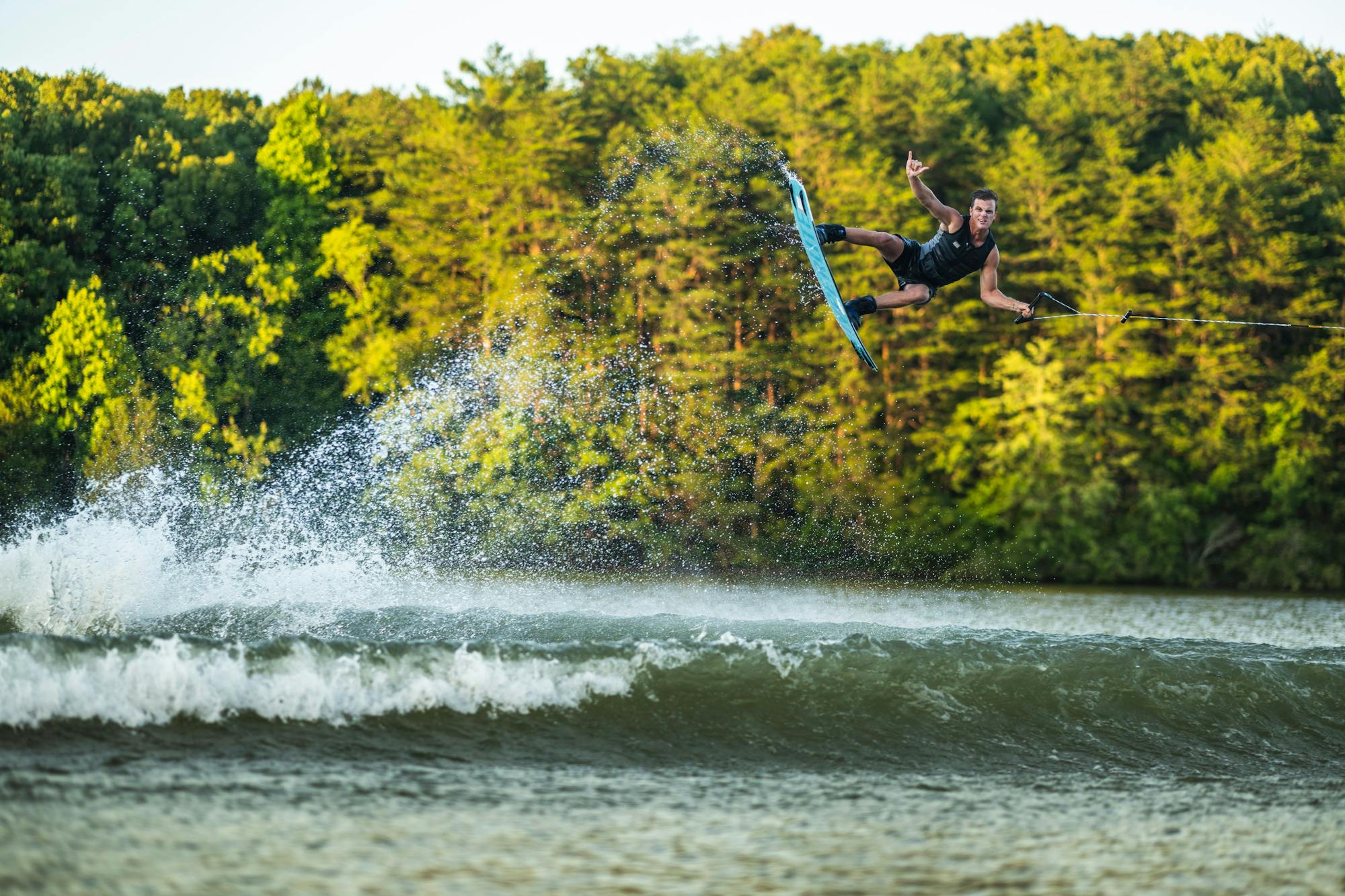 A person is wakeboarding on a lake, performing a trick in the air with one hand raised high. Their Hyperlite 2025 Sender Wakeboard, known for its sleek Dual Concave Base design, glides smoothly against the water. Forest trees stand tall in the background.