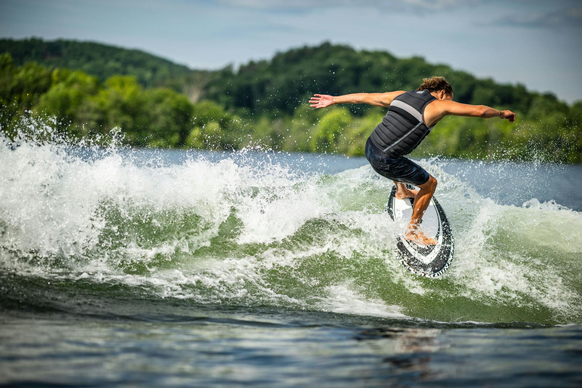 A person wakeboarding on a lake with a green forest backdrop, skillfully riding the waves on their Hyperlite 2025 Shim Wakesurf Board while wearing a vest crafted with DuraShell Construction.