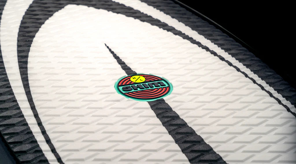 A detailed view of a textured surfboard displays the circular logo with the text "SHIM" and a smiling face. This Hyperlite 2025 Shim Wakesurf Board is crafted with DuraShell Construction and sports an eye-catching black and white pattern.