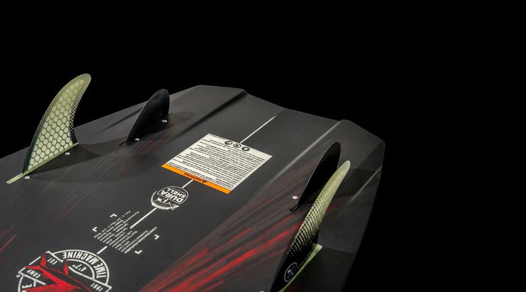 The image features the underside of the Hyperlite 2025 Time Machine Wakesurf Board, showcasing its three fins and surf style shape against a black background with graphic designs. A warning label is apparent, resembling a control panel on a time machine.