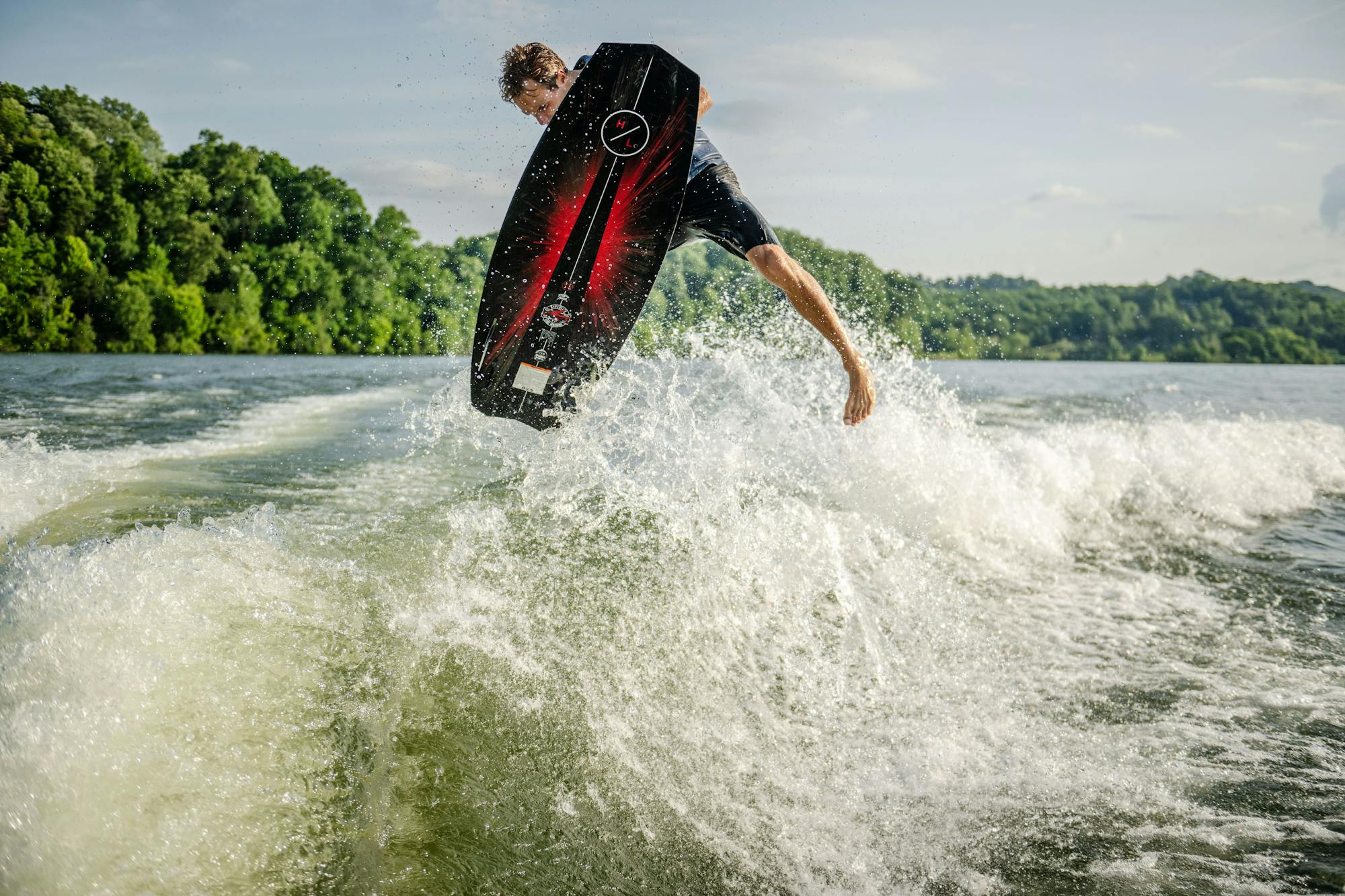 A person skillfully rides the Hyperlite 2025 Time Machine Wakesurf Board on a lake, performing an aerial maneuver with its surf style shape, surrounded by splashing water. The forested shoreline serves as a lush backdrop.