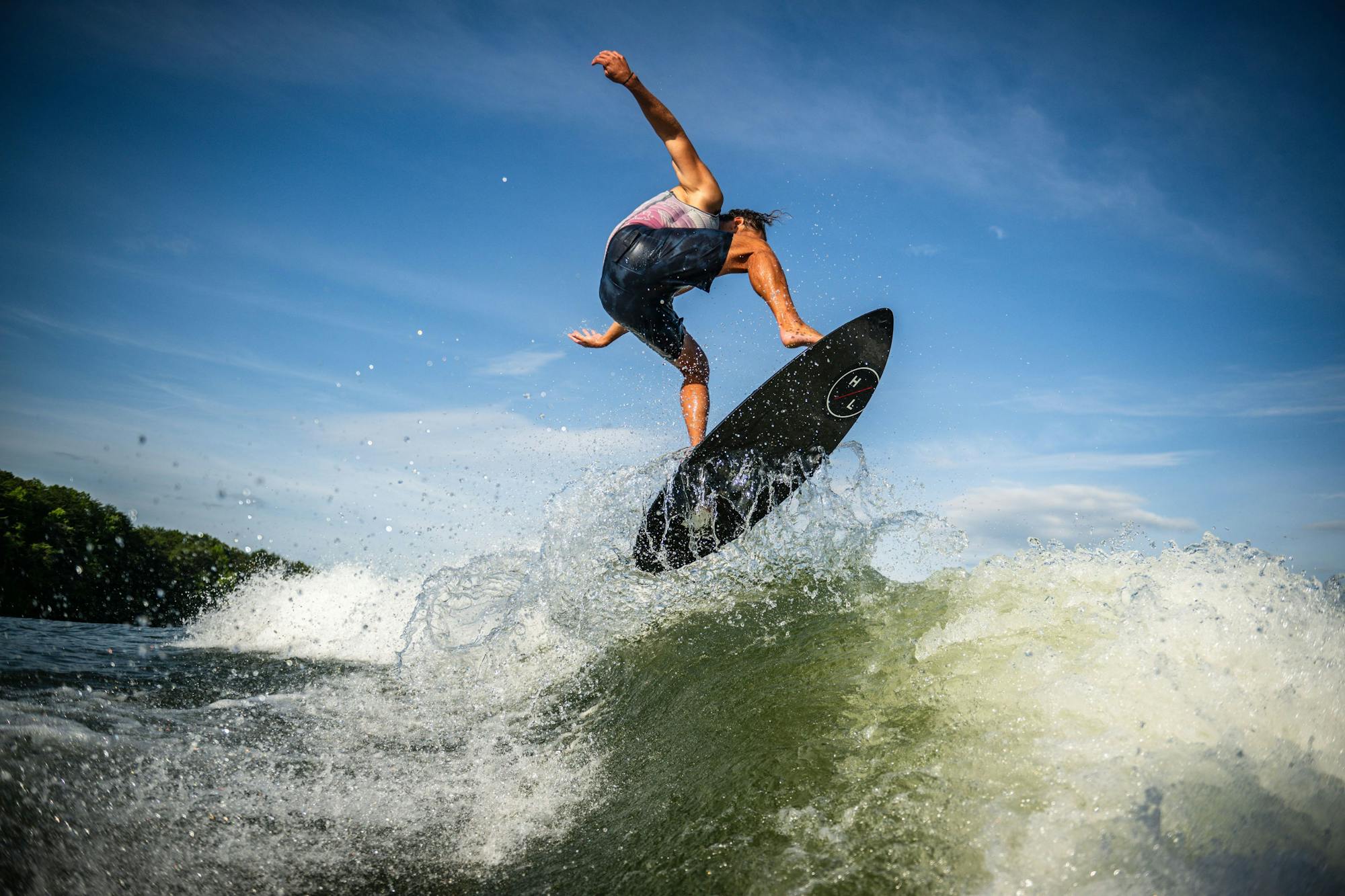 A person gracefully surfs on a wave, executing a mid-air maneuver against the clear blue sky, riding the cutting-edge Hyperlite 2025 Transistor Wakesurf Board.