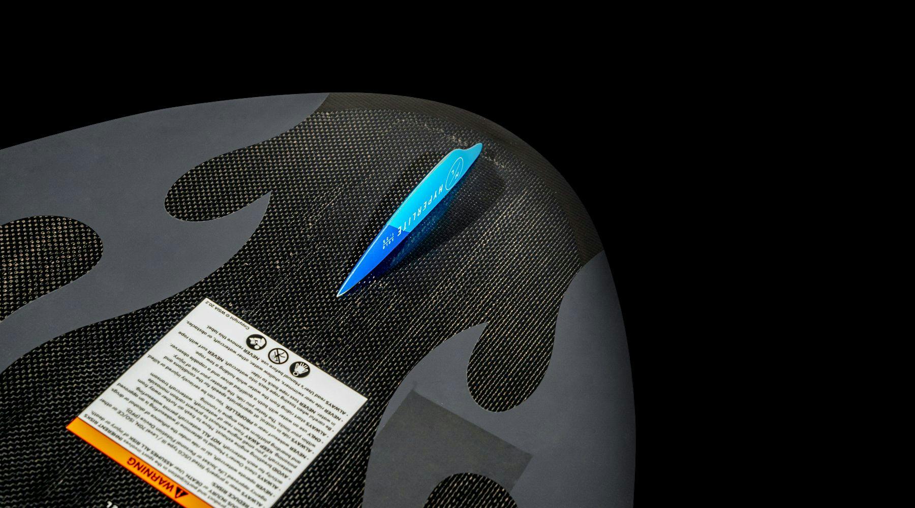 Close-up of the Hyperlite 2025 Transistor Wakesurf Board showcasing a black surface with flame-like patterns and a blue tag, featuring printed warnings and instructions, crafted with Nova Core woven carbon construction.